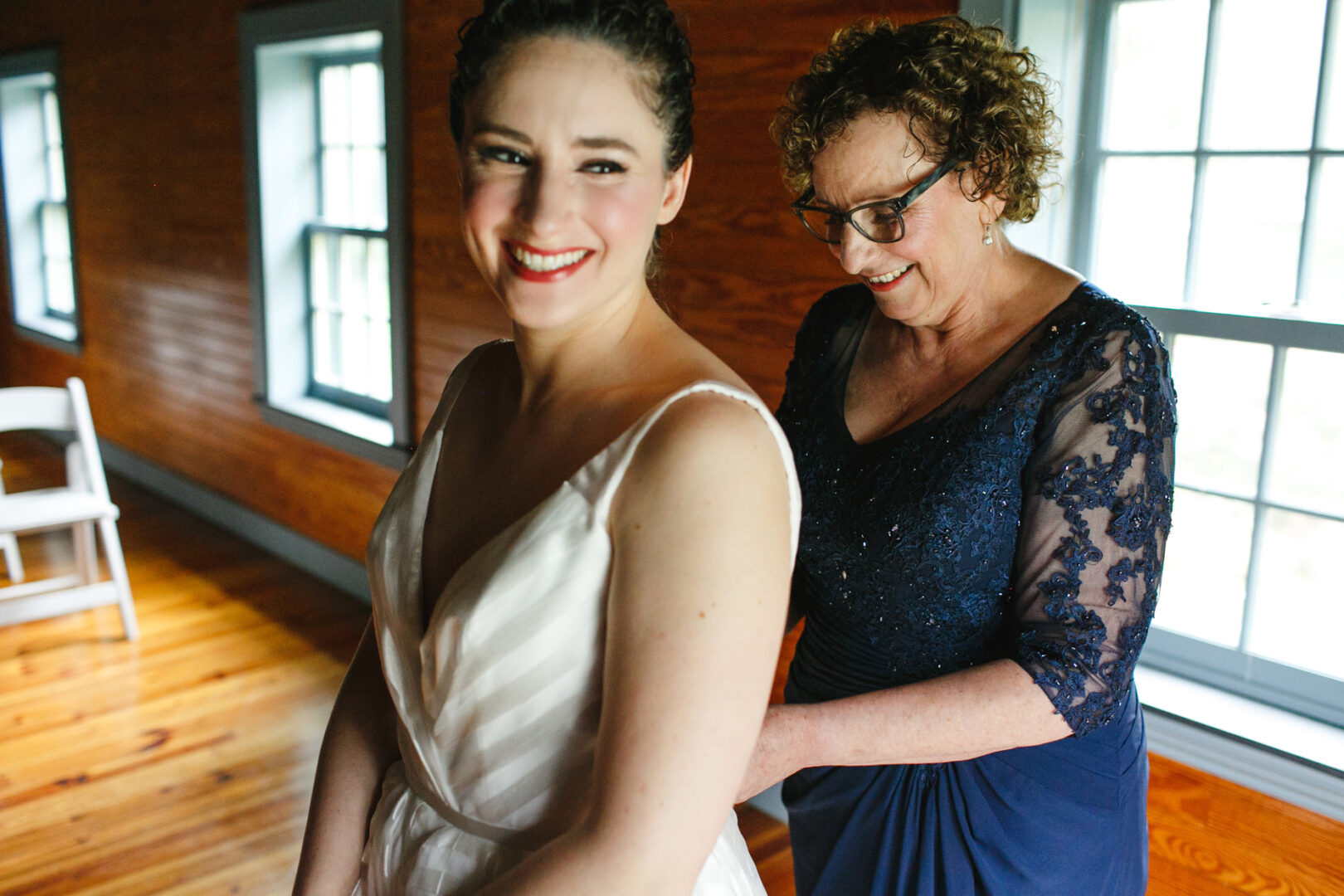 A bride in a white dress smiles as an older woman in navy helps with the back, bathed in sunlight streaming through the wooden walls of Rockwood Park Carriage House.