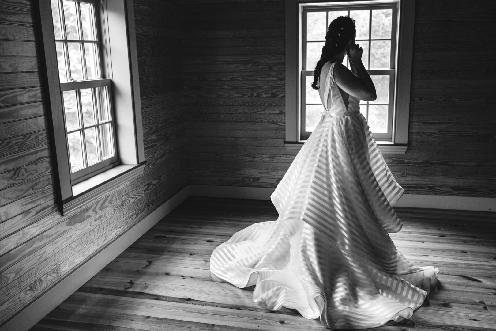 A woman in a flowing wedding gown stands in the wooden elegance of Rockwood Park Carriage House, gazing thoughtfully out of a window.