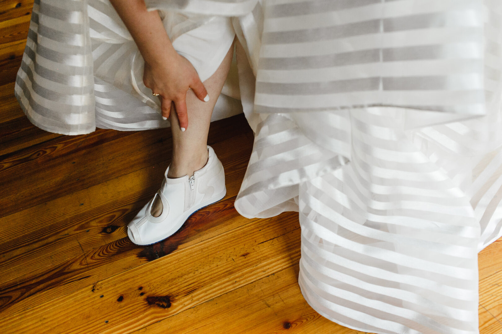 Person in a white striped dress adjusts their left leg while wearing white sneakers on the wooden floor of Rockwood Park Carriage House, capturing a moment both casual and elegant.
