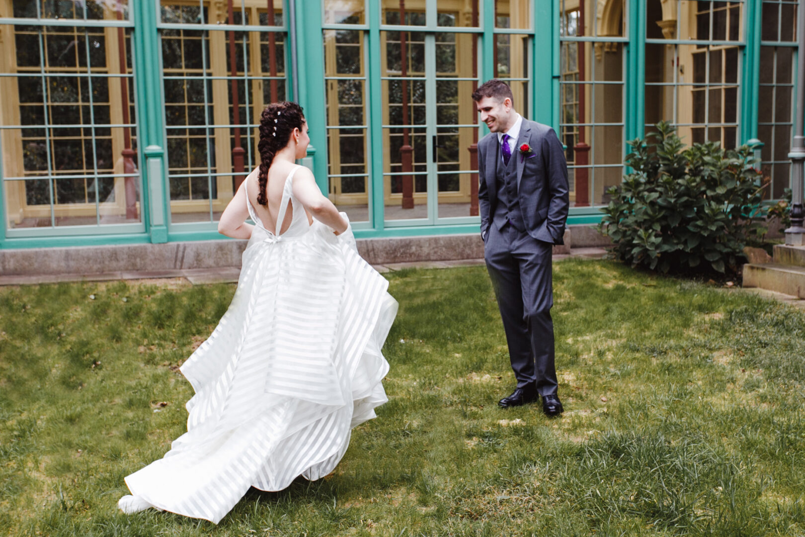 The bride, in a striped gown, and the groom, sporting a purple boutonniere, stand elegantly on the lush grass near the vibrant green and yellow Rockwood Park Carriage House with its grand windows.