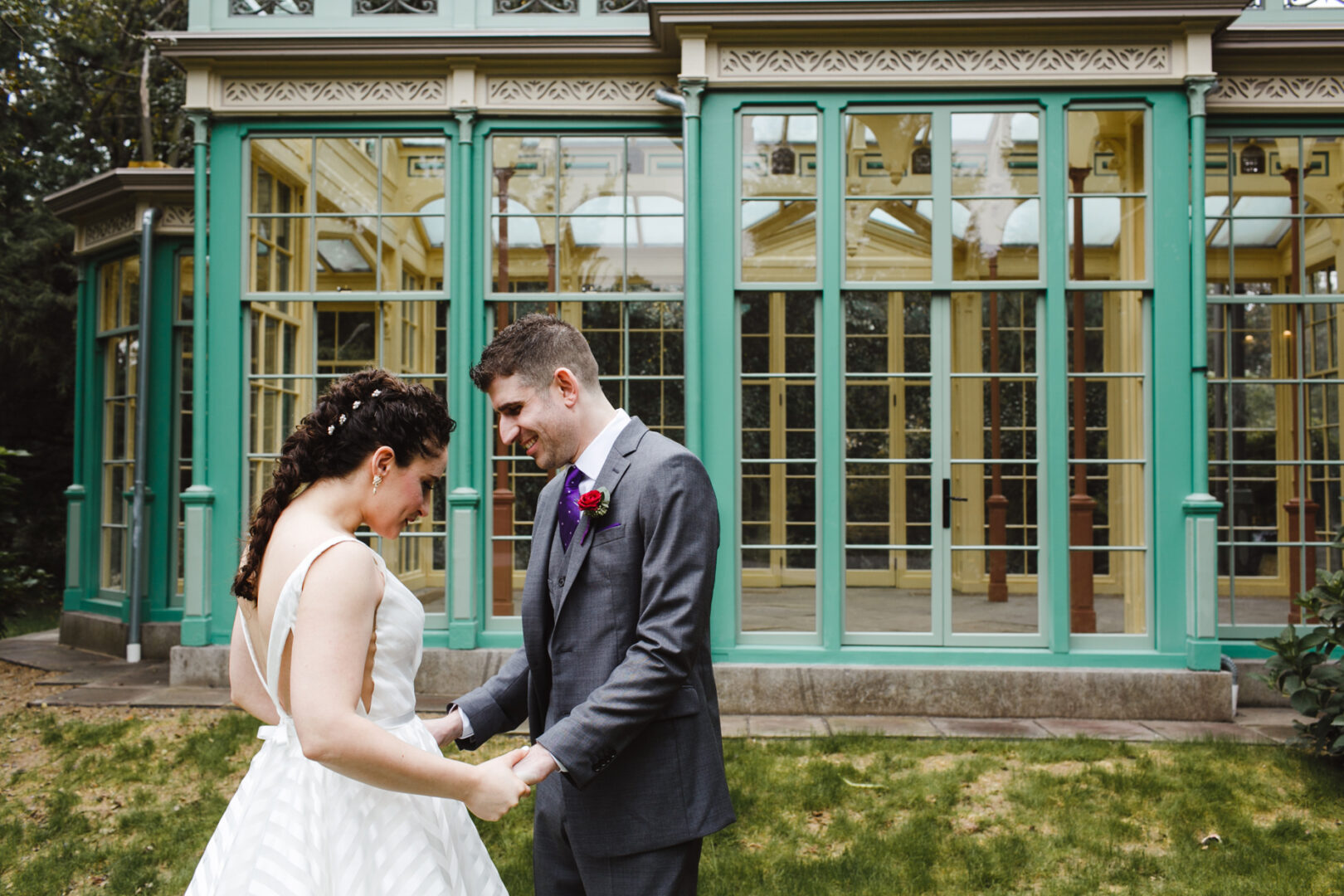 A smiling couple in wedding attire stands holding hands on a grassy area before the Rockwood Park Carriage House, its large glass and green-framed structure creating a stunning backdrop.