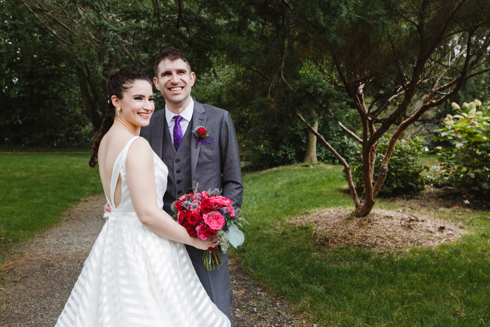 A couple in wedding attire smiles while standing on a path in a wooded area at their Rockwood Park Carriage House wedding. The bride holds a bouquet of red flowers, capturing the perfect blend of natural beauty and elegant charm.