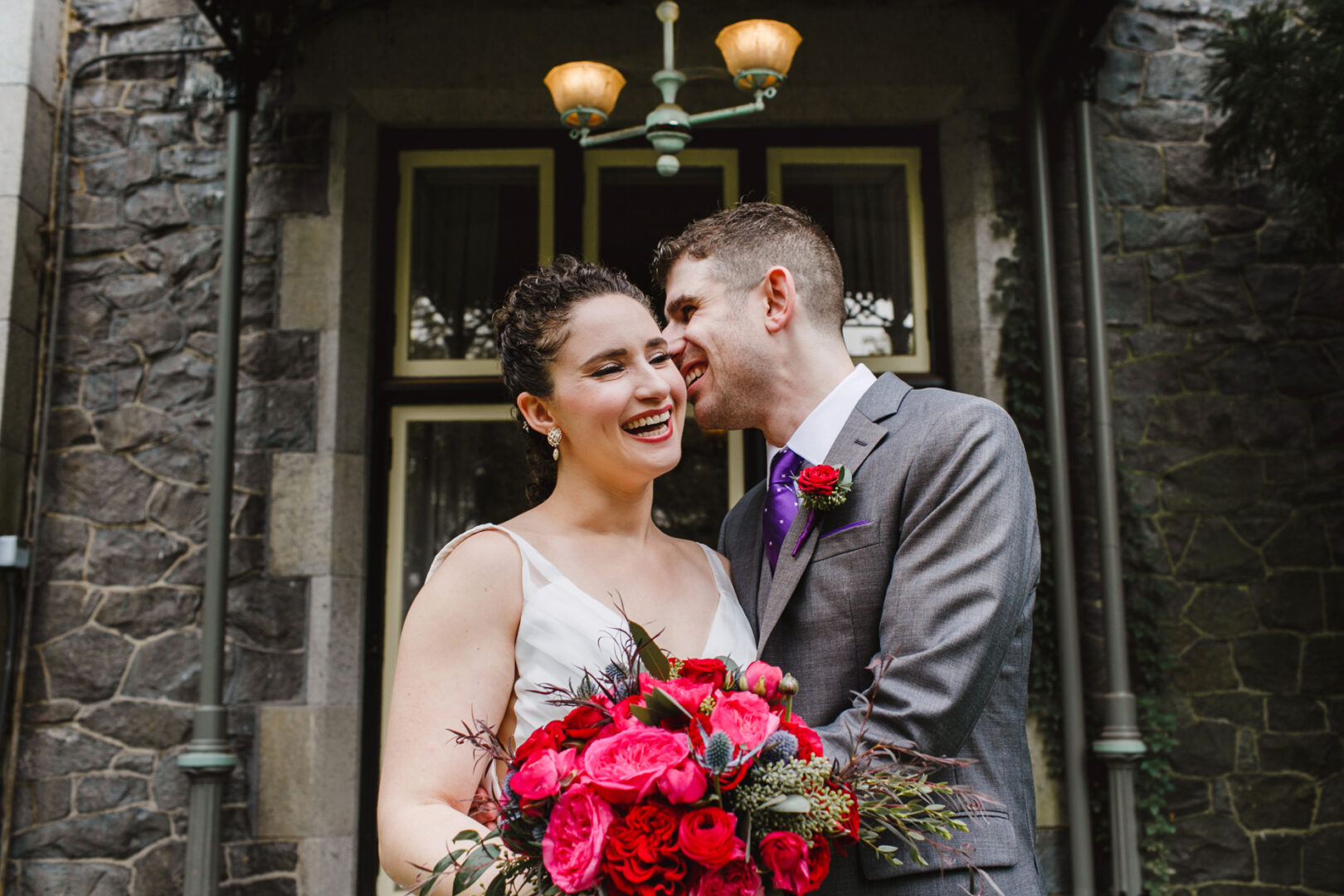 A couple in formal attire stands outside the historic Rockwood Park Carriage House. The man leans in to whisper to the smiling woman holding a vibrant bouquet of red flowers, capturing a moment of elegance and romance on their wedding day.