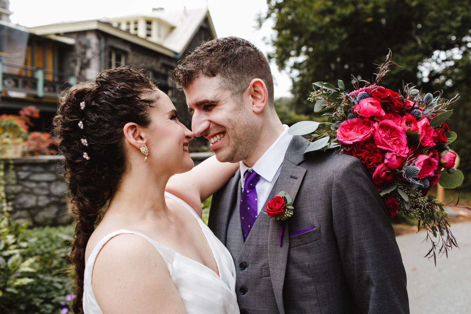 At their Rockwood Park Carriage House wedding, the bride and groom smile at each other outdoors. The bride holds a bouquet of red and pink flowers, adding a vibrant touch to the enchanting scene.