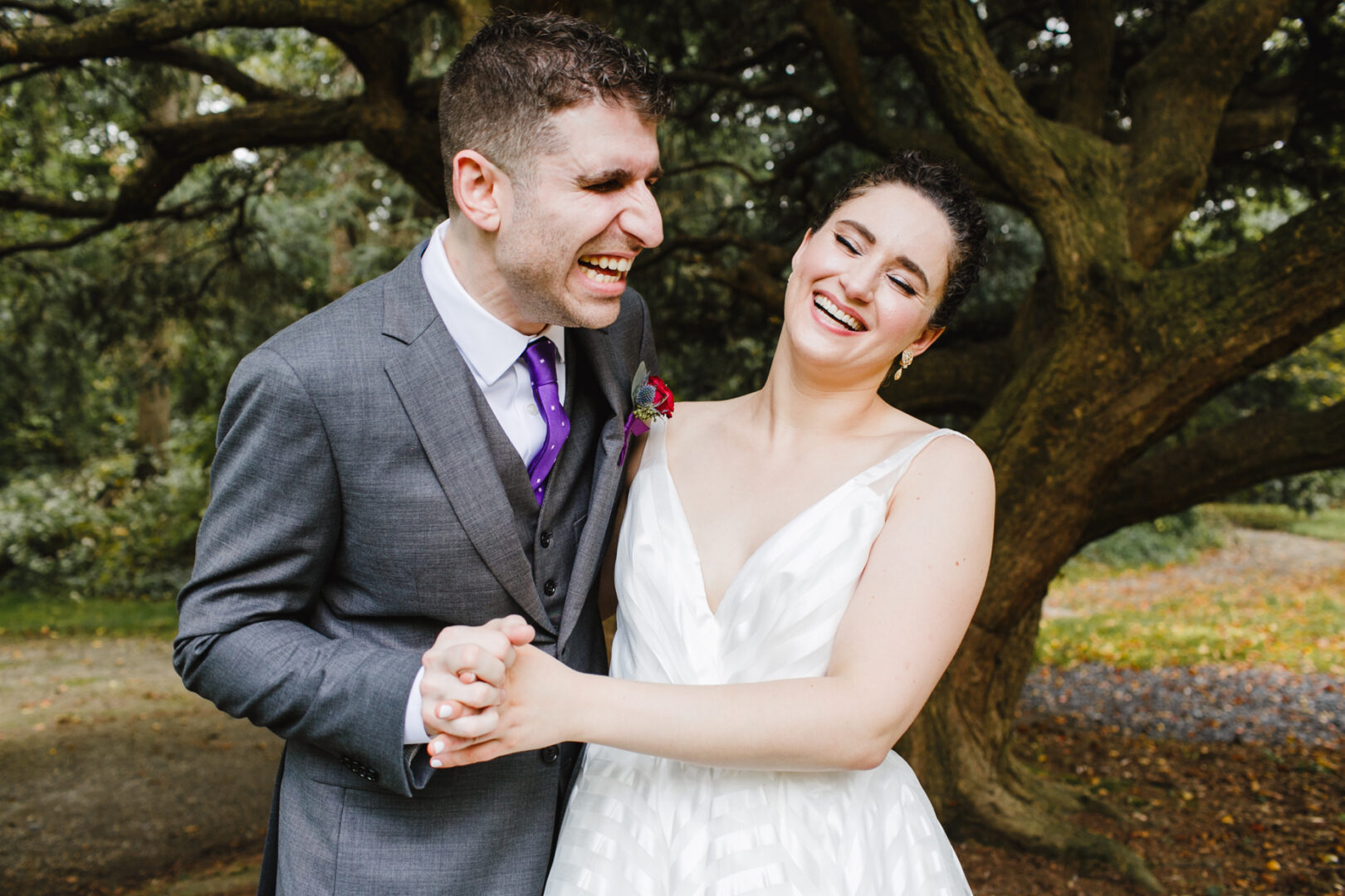 A smiling couple, with one in a gray suit and the other in a white dress, stand hand in hand outdoors by a large tree, capturing their joy on a special day at the Rockwood Park Carriage House Wedding.