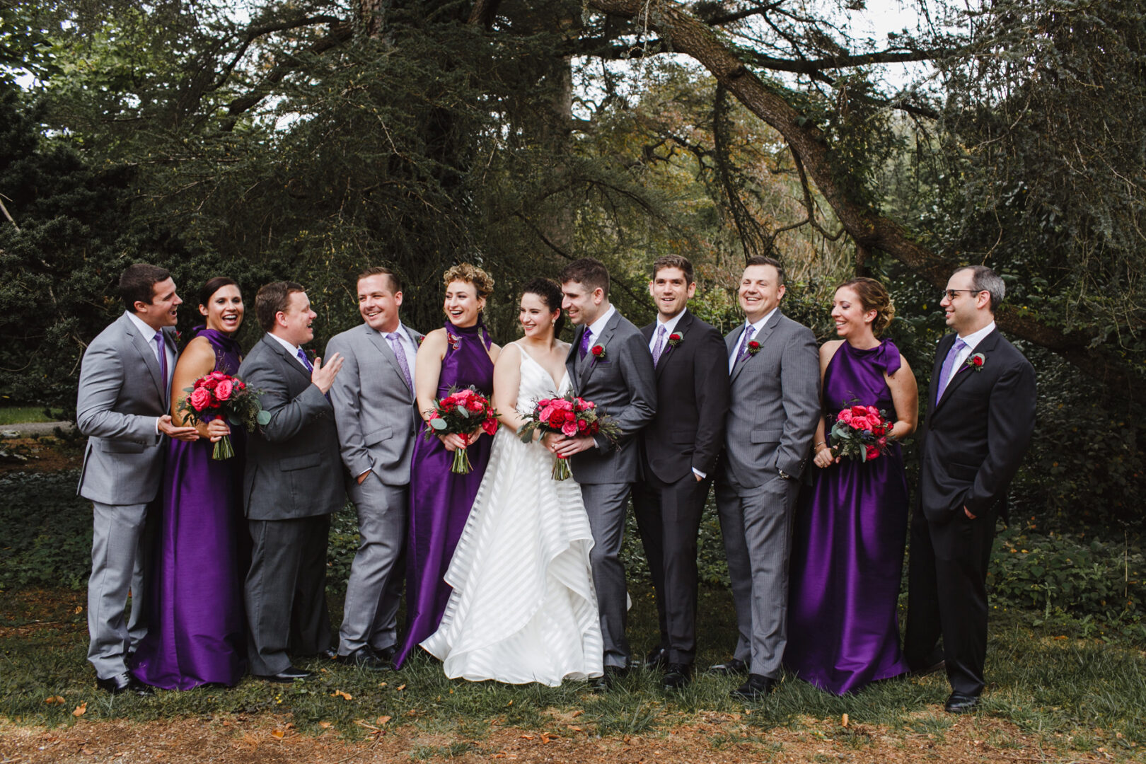 A wedding party stands outdoors at Rockwood Park Carriage House, with the bride and groom in the center. Bridesmaids in purple dresses hold red bouquets; groomsmen wear gray suits. Trees and greenery provide a stunning backdrop.