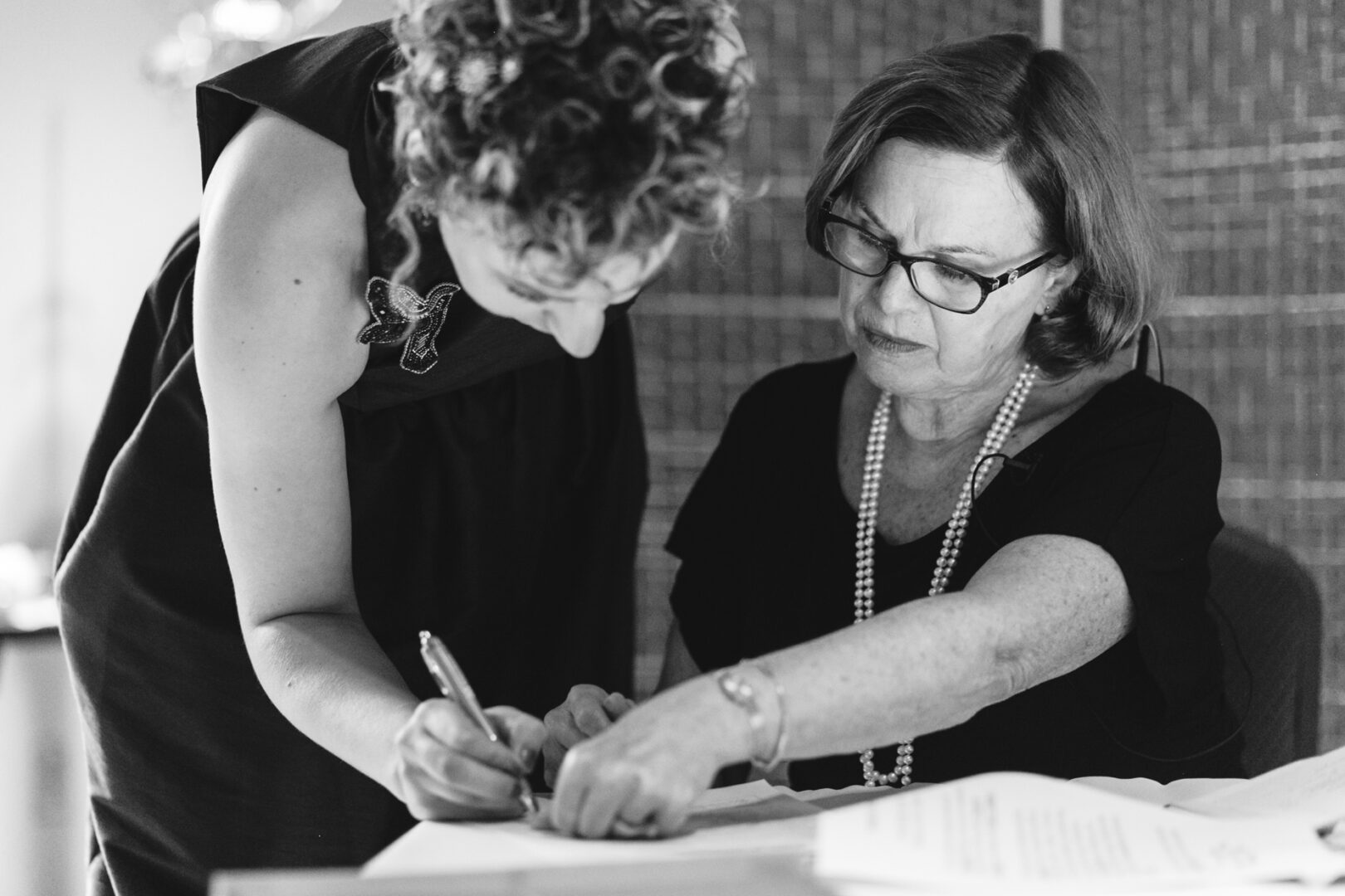 In the elegant setting of Rockwood Park Carriage House, two women are concentrated on paperwork. One stands, writing intently, while the other sits, pointing at the document. Both are dressed formally, embodying a blend of professionalism and refined charm.