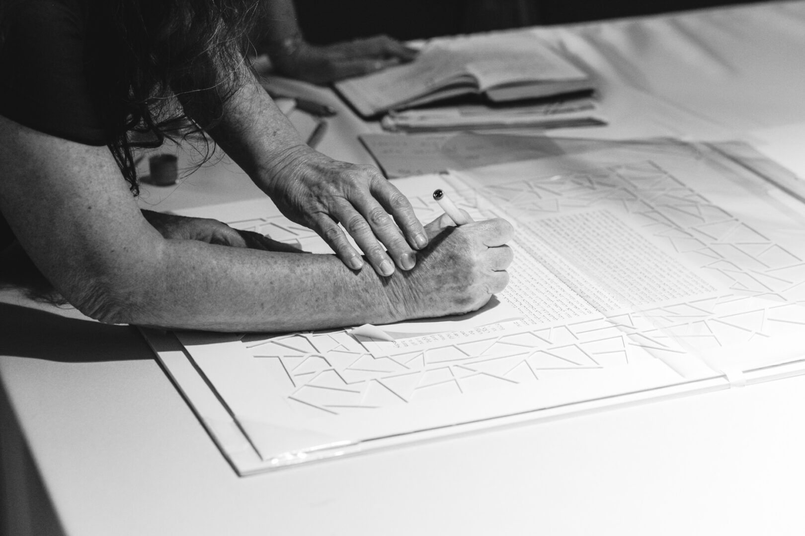 A person meticulously writing on a sheet of paper adorned with geometric patterns sits at a table, where an open notebook lies beside a closed book, perhaps planning the perfect Rockwood Park Carriage House wedding.