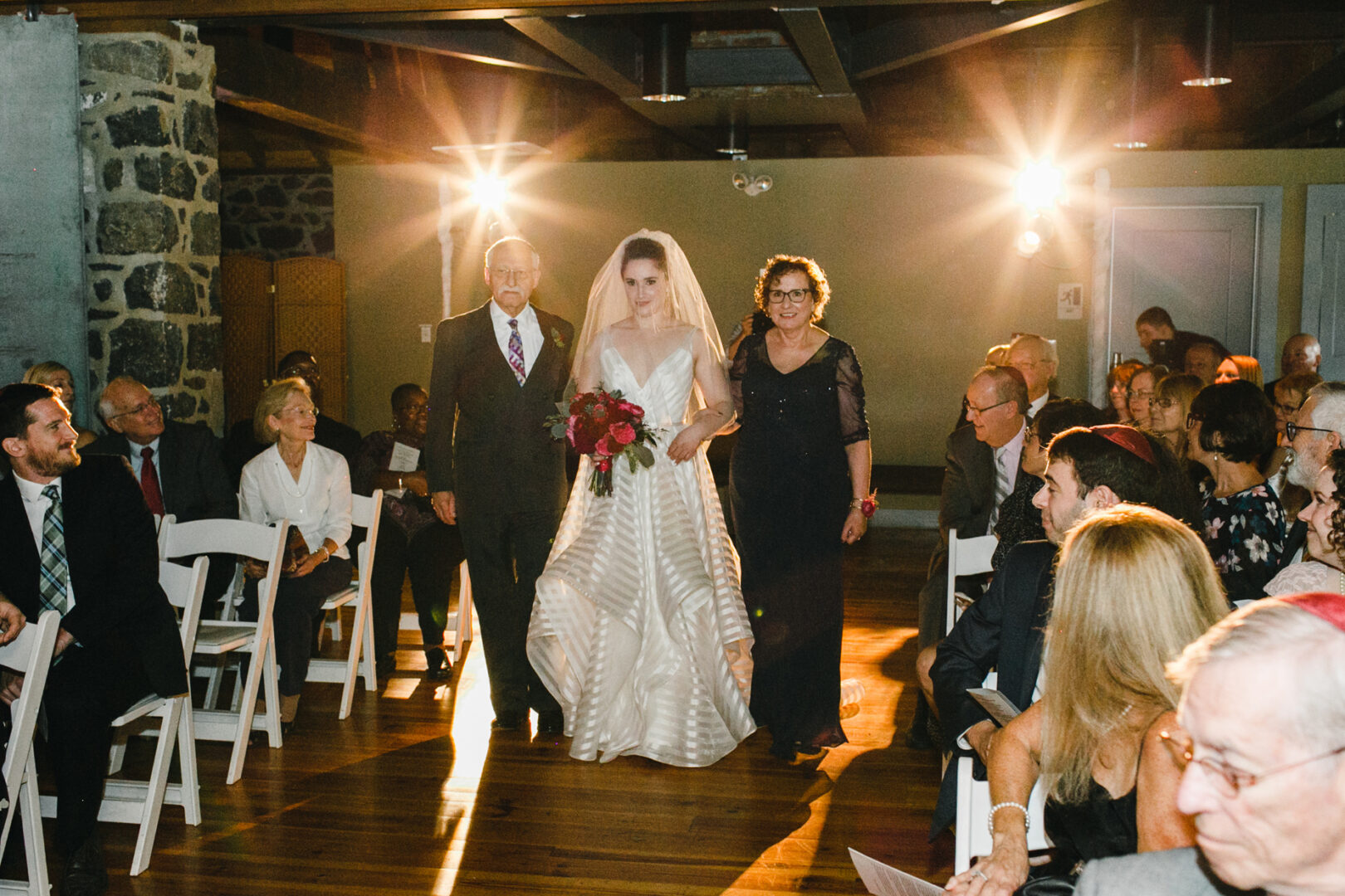A bride in a white dress walks down the aisle at Rockwood Park Carriage House, accompanied by an older man and woman, as guests seated on either side look on in a dimly lit room.