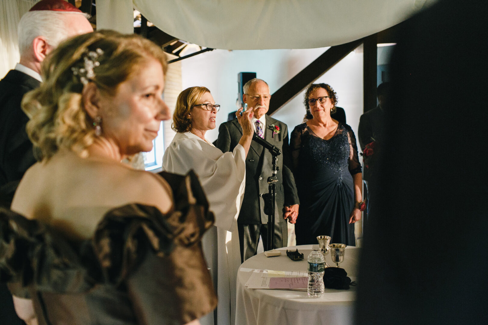 A wedding ceremony unfolds at the scenic Rockwood Park Carriage House. Indoors, a person in a white robe speaks into a microphone as the couple, hands clasped, faces them. Other attendees are partially visible, adding to the intimate ambiance.