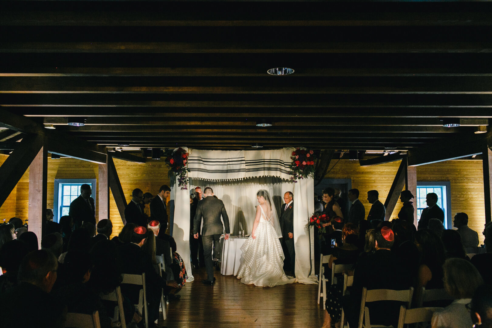 A Rockwood Park Carriage House wedding unfolds with a couple under a canopy, accompanied by two people. Guests are seated on either side in the dimly lit room, adorned with elegant wooden beams.