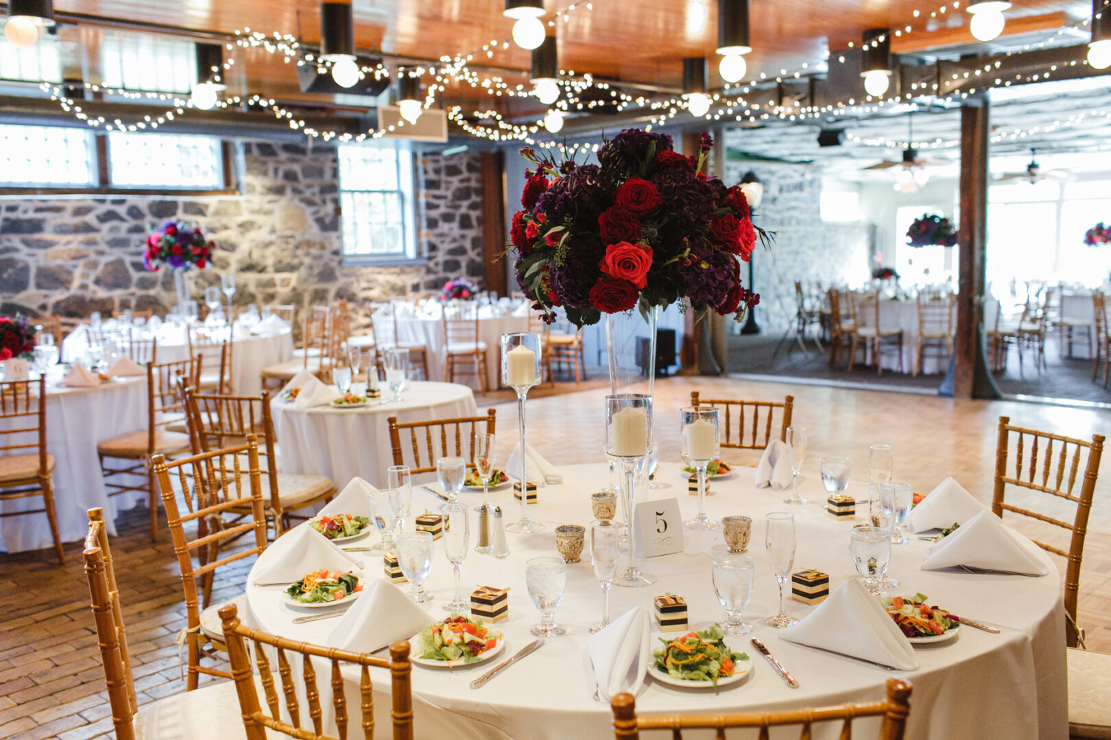 The Rockwood Park Carriage House Wedding featured a beautifully decorated banquet hall with round tables set for the event, showcasing white tablecloths, salads, desserts, candles, and floral centerpieces all elegantly illuminated under string lights.