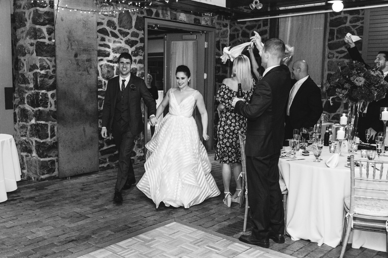A couple in formal attire enters the Rockwood Park Carriage House Wedding venue, greeted by standing guests with napkins raised. The setting features brick and stone walls, with string lights twinkling above.