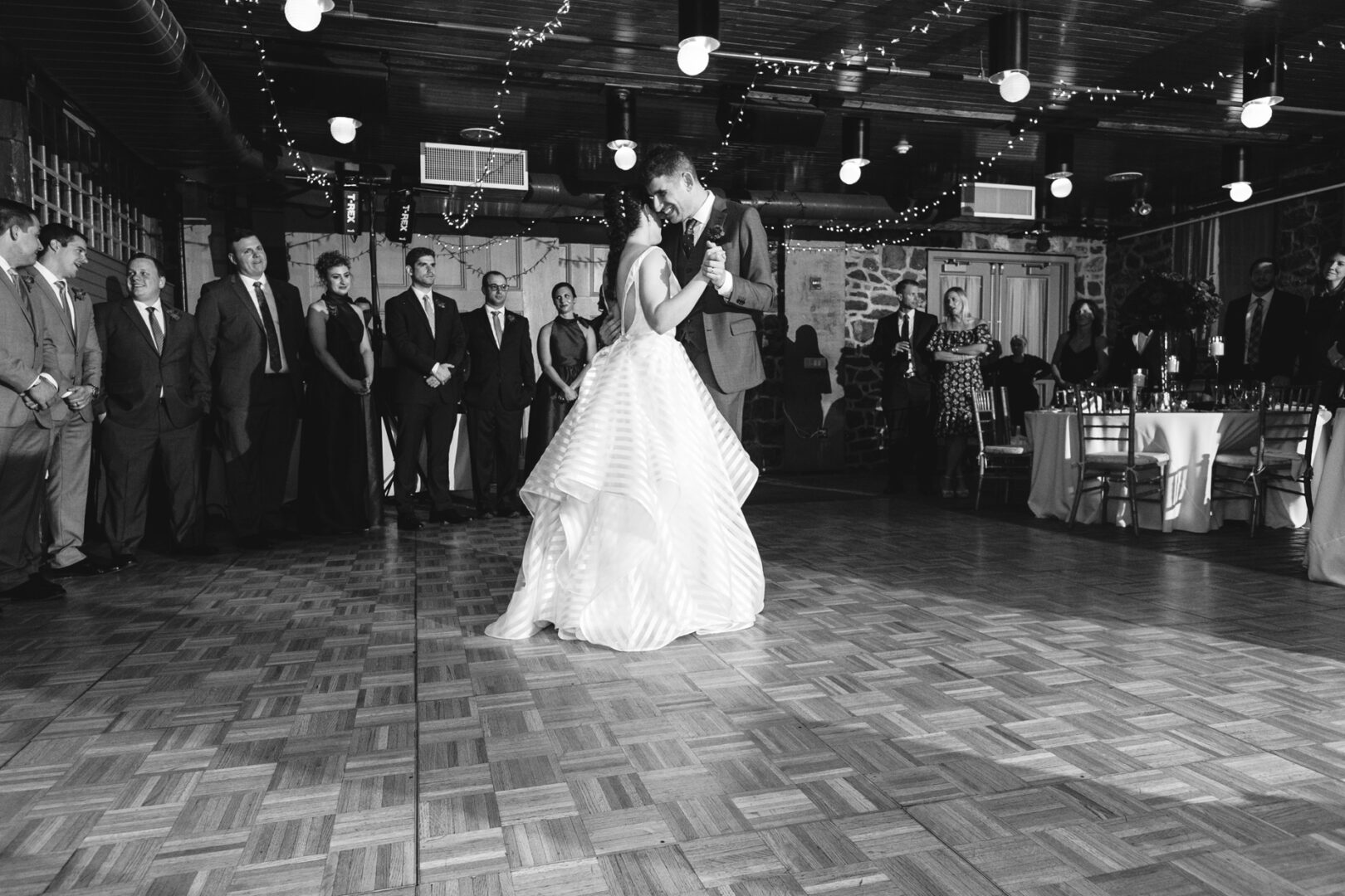 At their Rockwood Park Carriage House wedding, a couple dances gracefully on a wooden floor, surrounded by guests in formal attire as string lights twinkle overhead.