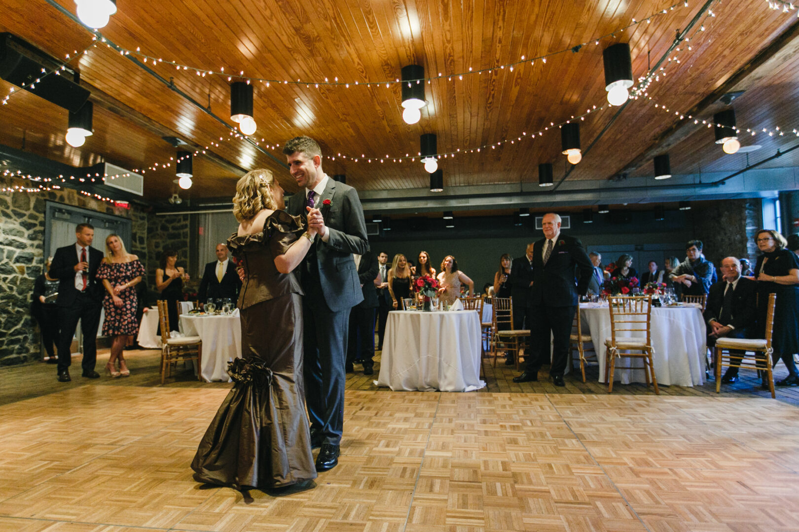 A couple gracefully dances on the wooden floor of the Rockwood Park Carriage House, surrounded by seated and standing guests in a beautifully decorated event hall.