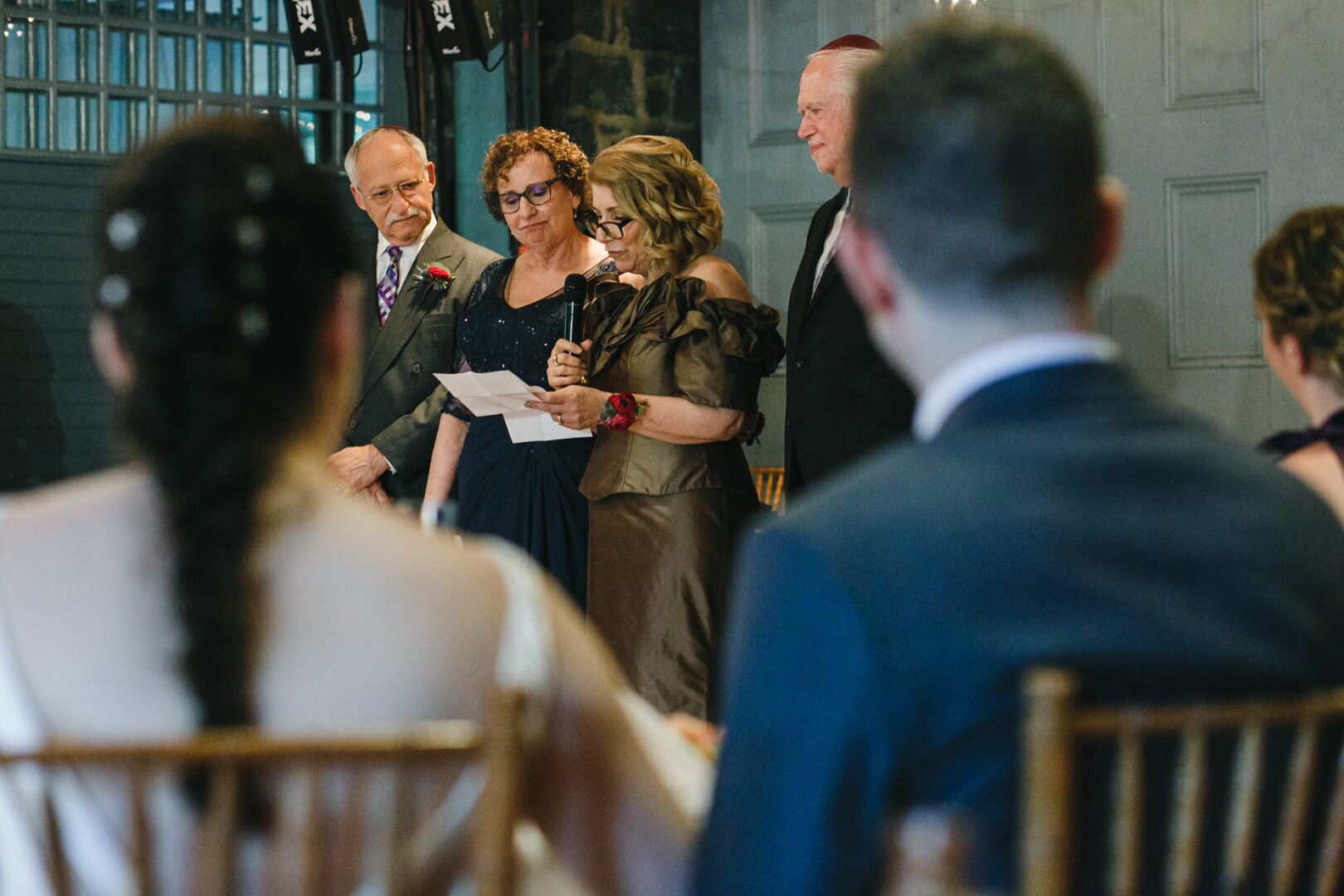 At the Rockwood Park Carriage House wedding, a group gathered indoors for the formal event. One woman read from a paper as others watched intently. In the foreground, two more guests were seated, slightly out of focus, quietly absorbing the moment.