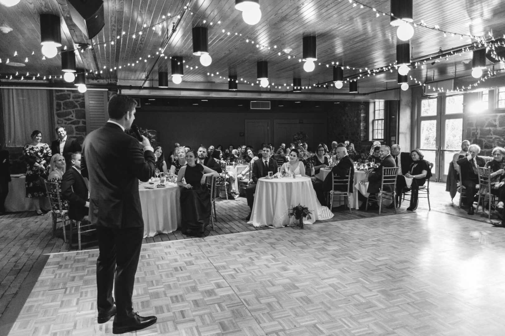 At the Rockwood Park Carriage House Wedding, a man delivers a speech in a beautifully decorated hall. Guests sit at round tables, listening intently as string lights twinkle above, illuminating the elegant wooden flooring.