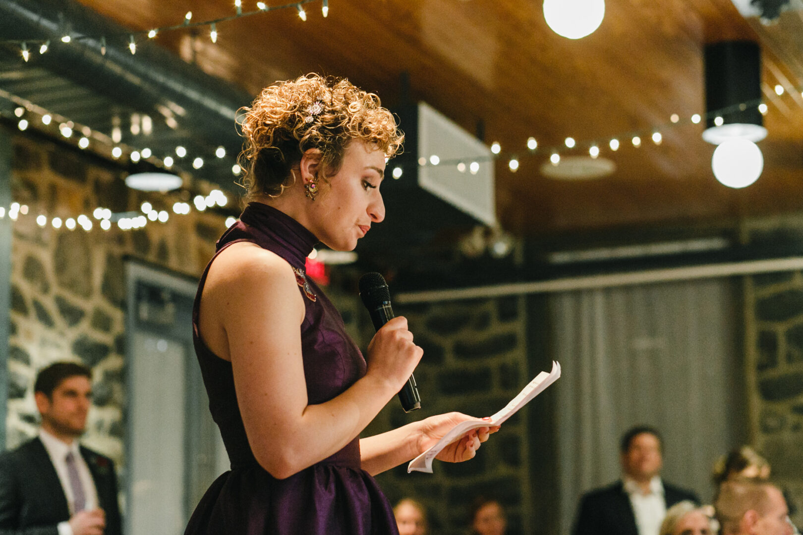 In the warmly lit Rockwood Park Carriage House, a person in a purple dress speaks into a microphone while holding a piece of paper, surrounded by the gentle glow of string lights—a true wedding moment to cherish.