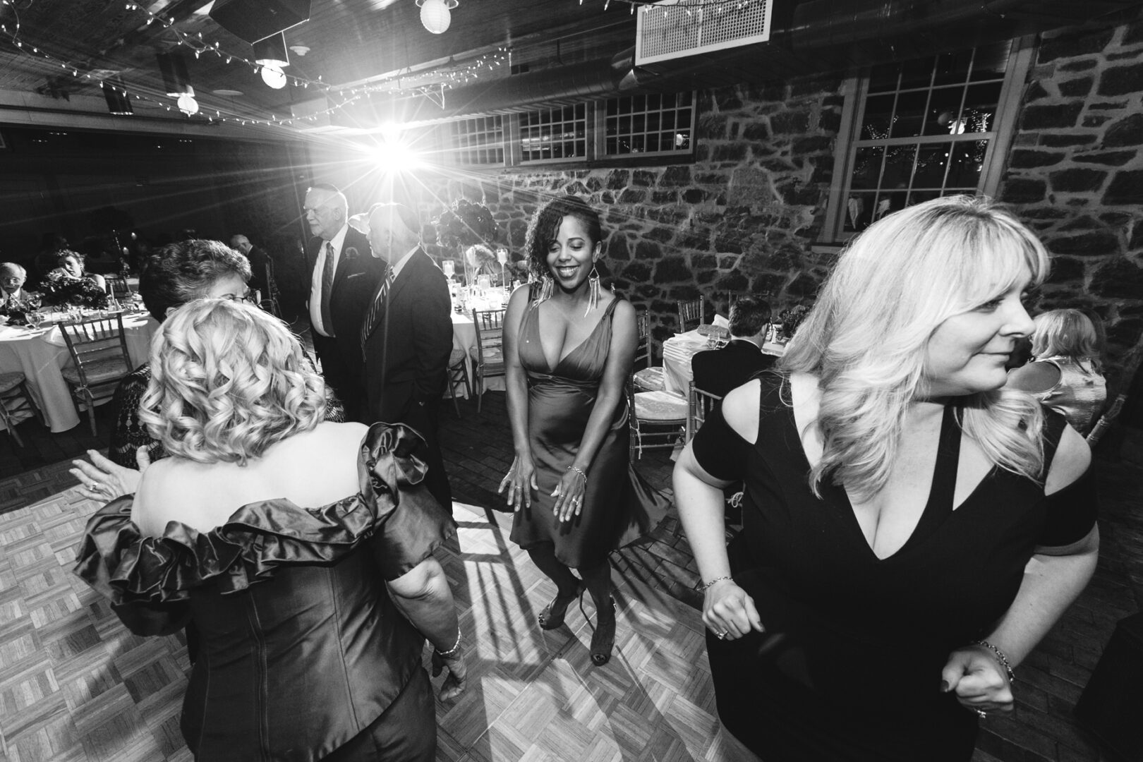 A group of people dressed in formal attire are dancing energetically on a wooden floor at the Rockwood Park Carriage House Wedding, with dim lighting and a bright light shining from the background.