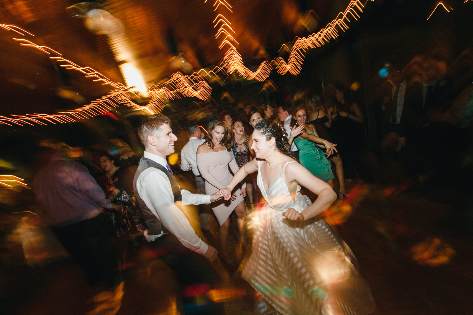 Amidst the vibrant atmosphere of a Rockwood Park Carriage House wedding, a couple dances energetically at the lively event, surrounded by an exuberant crowd. Blurred lights enhance the festive mood as everyone revels in the joy of the celebration.