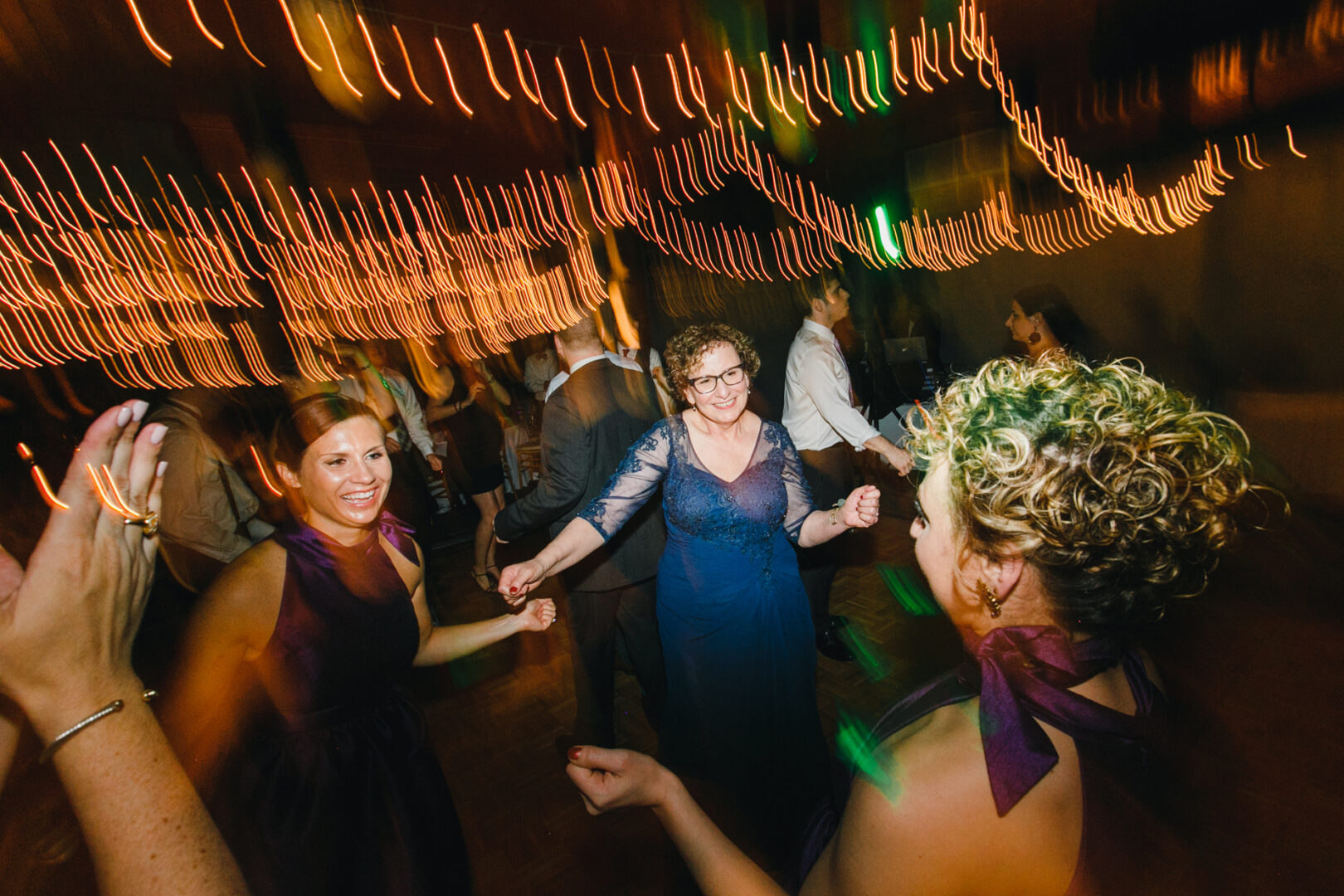 People dancing in a dimly lit venue at the Rockwood Park Carriage House Wedding, with string lights creating motion blur. Three women are visibly enjoying the dance, surrounded by others.