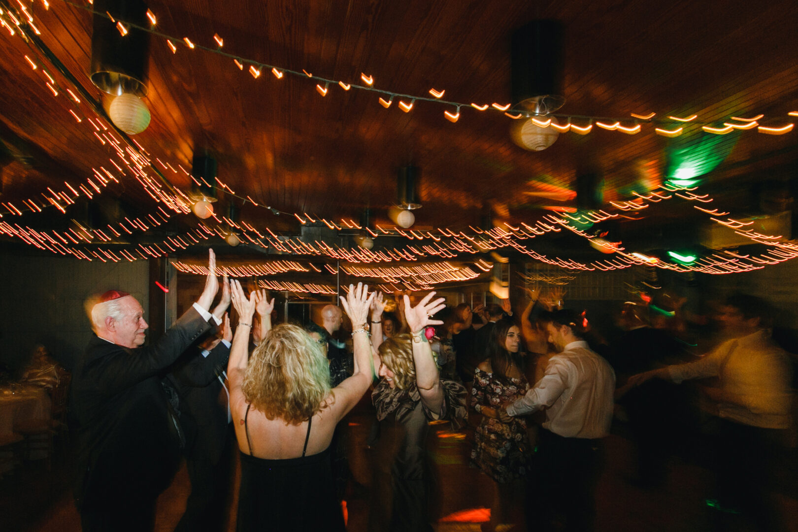 People dancing under warm string lights at an indoor celebration in the Rockwood Park Carriage House, with a wooden ceiling and blurred motion capturing their movements.