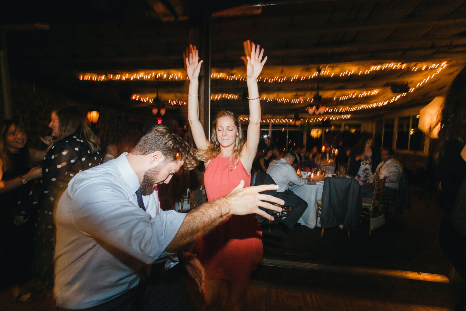 Two people dancing enthusiastically at a dimly lit Rockwood Park Carriage House wedding, with string lights and tables in the background.