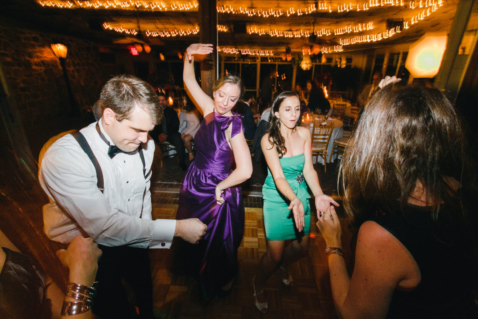 At the Rockwood Park Carriage House wedding, people are dancing enthusiastically under string lights. One person sports a white shirt with suspenders, while two others twirl in vibrant purple and green dresses, adding to the joyous celebration.