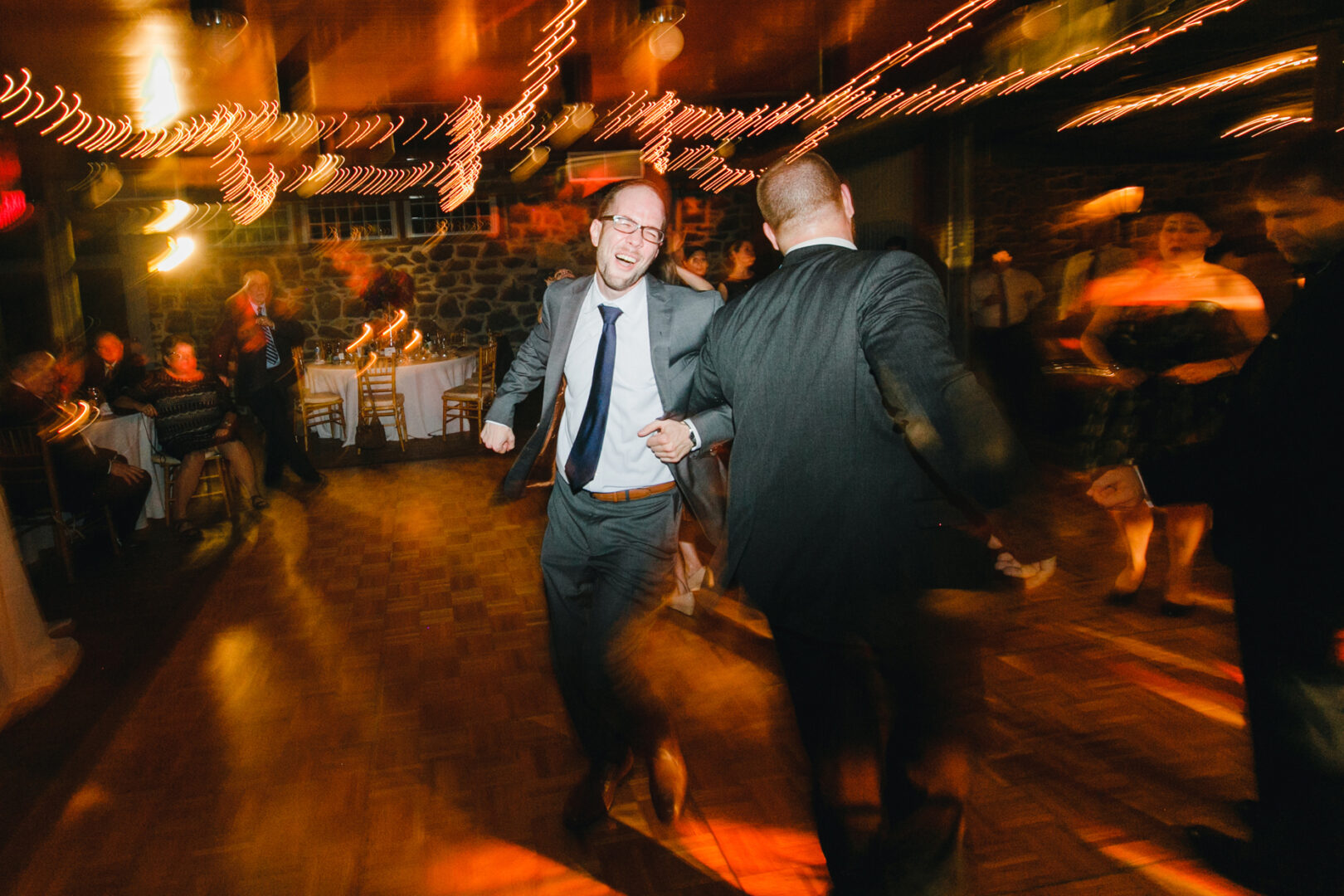 Men in suits dance energetically on the wooden floor of Rockwood Park Carriage House, surrounded by blurred figures and string lights, creating a magical ambiance in the dimly lit room.