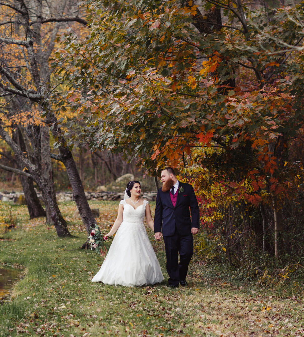 A bride in a white dress and a groom in a black suit walk hand in hand along a tree-lined path by the lake at Brandywine Manor House, surrounded by the vibrant autumn foliage of their wedding day.