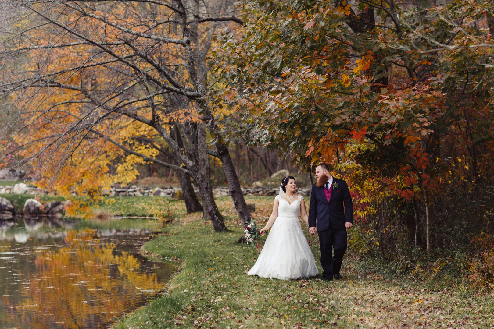 A bride in a white dress and a groom in a black suit walk hand in hand along a tree-lined path by the lake at Brandywine Manor House, surrounded by the vibrant autumn foliage of their wedding day.