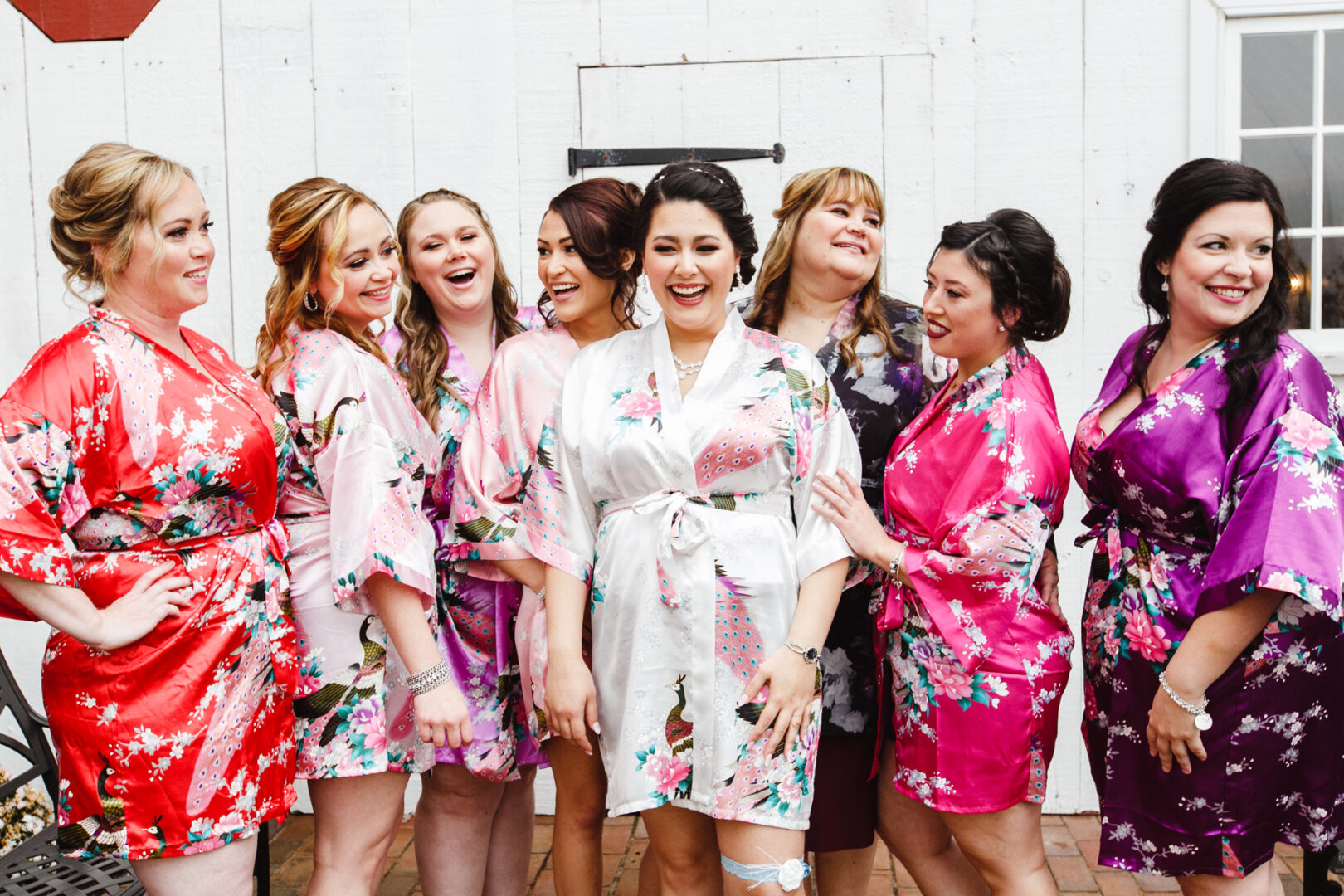 A group of women in floral robes smile and laugh together outdoors, capturing the joy of a Brandywine Manor House wedding in front of a charming white wall.