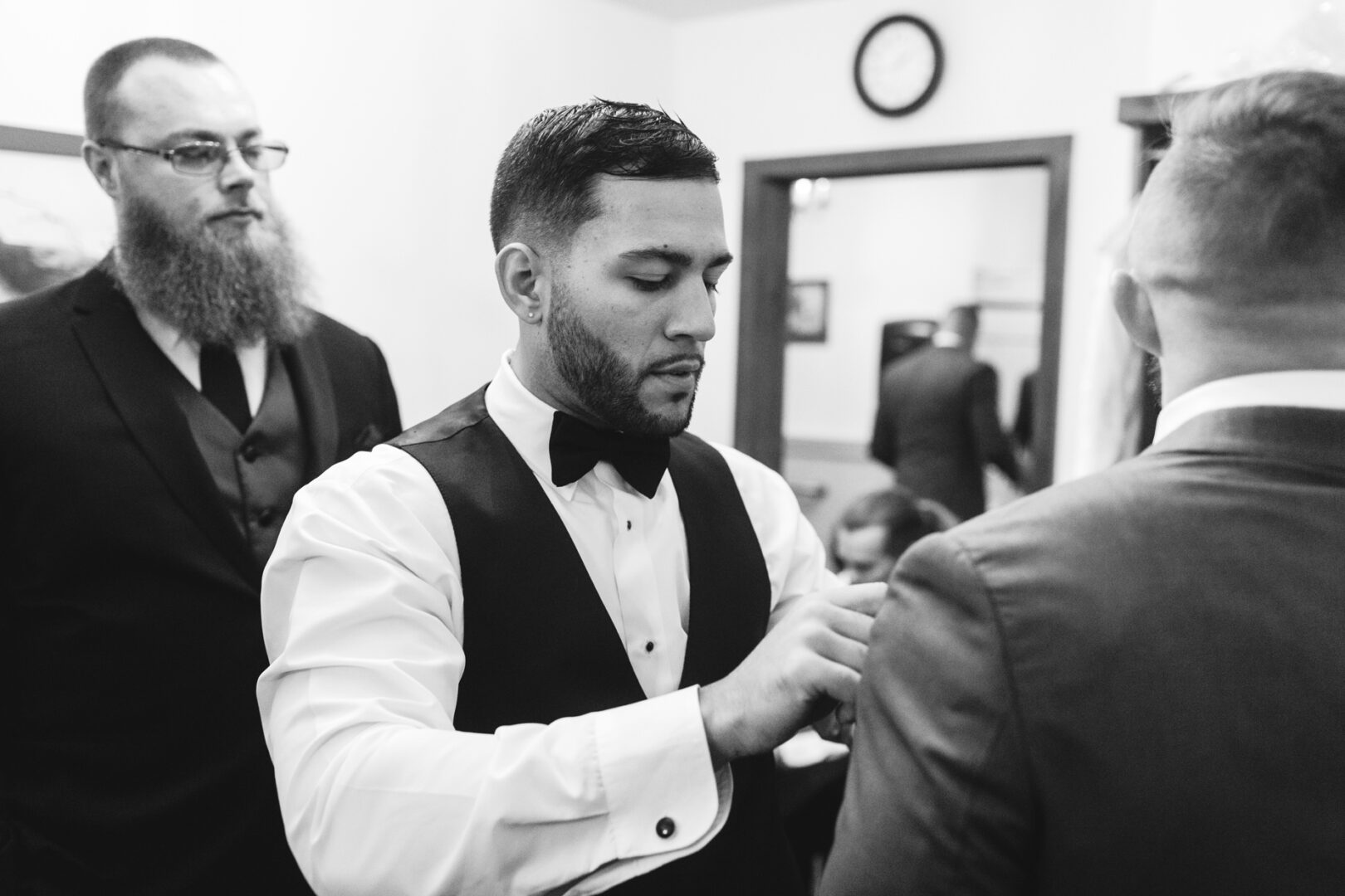 In a room at Brandywine Manor House, a man in a tuxedo adjusts another man's suit while a third observes with interest. A clock on the wall quietly marks the time as they prepare for the wedding, all impeccably dressed in formal attire.