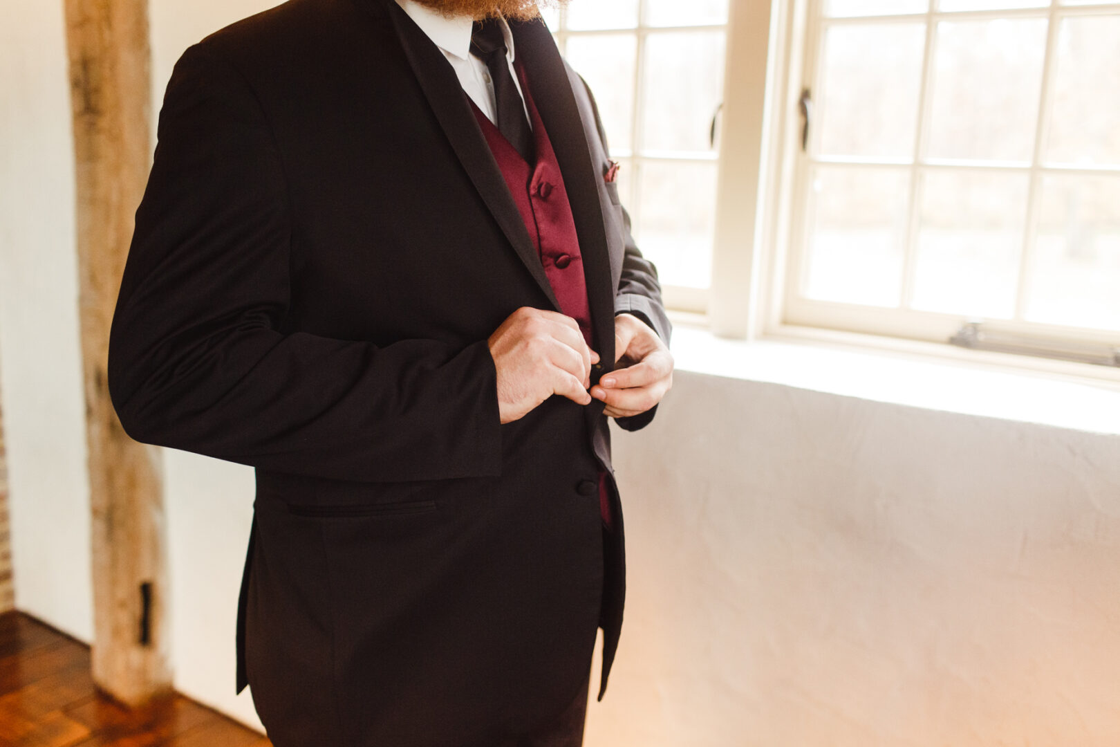 In a room with large windows, a person in a dark suit and red vest adjusts their jacket, preparing for a memorable Brandywine Manor House wedding.