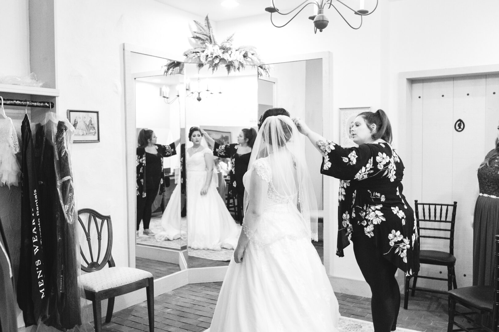 A bride stands in front of a mirror at Brandywine Manor House Wedding as a woman adjusts her veil in the dressing room. Two chairs and hanging garments complete the serene scene.