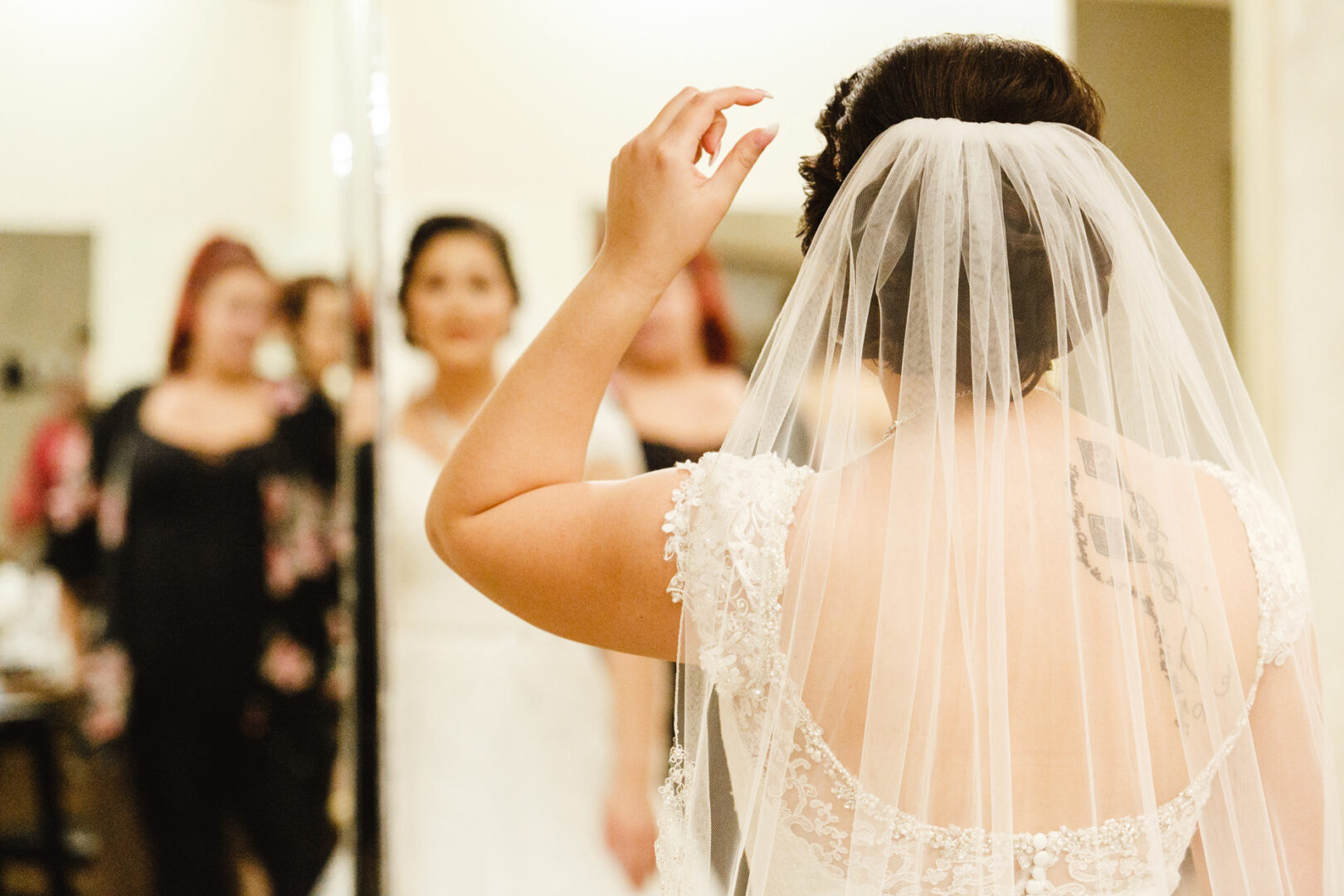 In the serene ambiance of Brandywine Manor House, a bride with a veil stands gracefully before the mirror, adjusting her hair as women in elegant attire gather in the background.