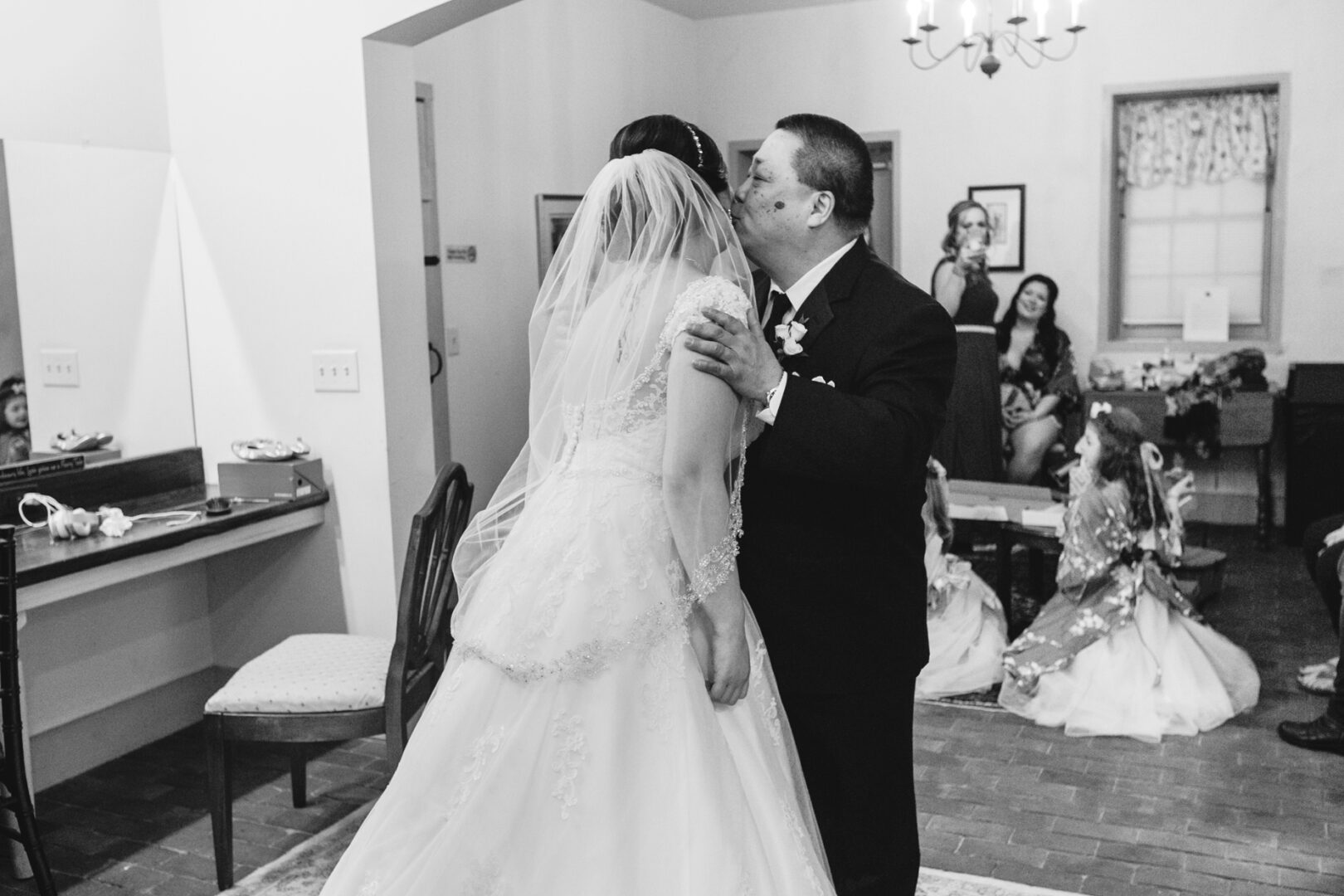 In the elegant setting of Brandywine Manor House, a man tenderly kisses the bride on the cheek, while bridesmaids gather nearby in joyful anticipation.