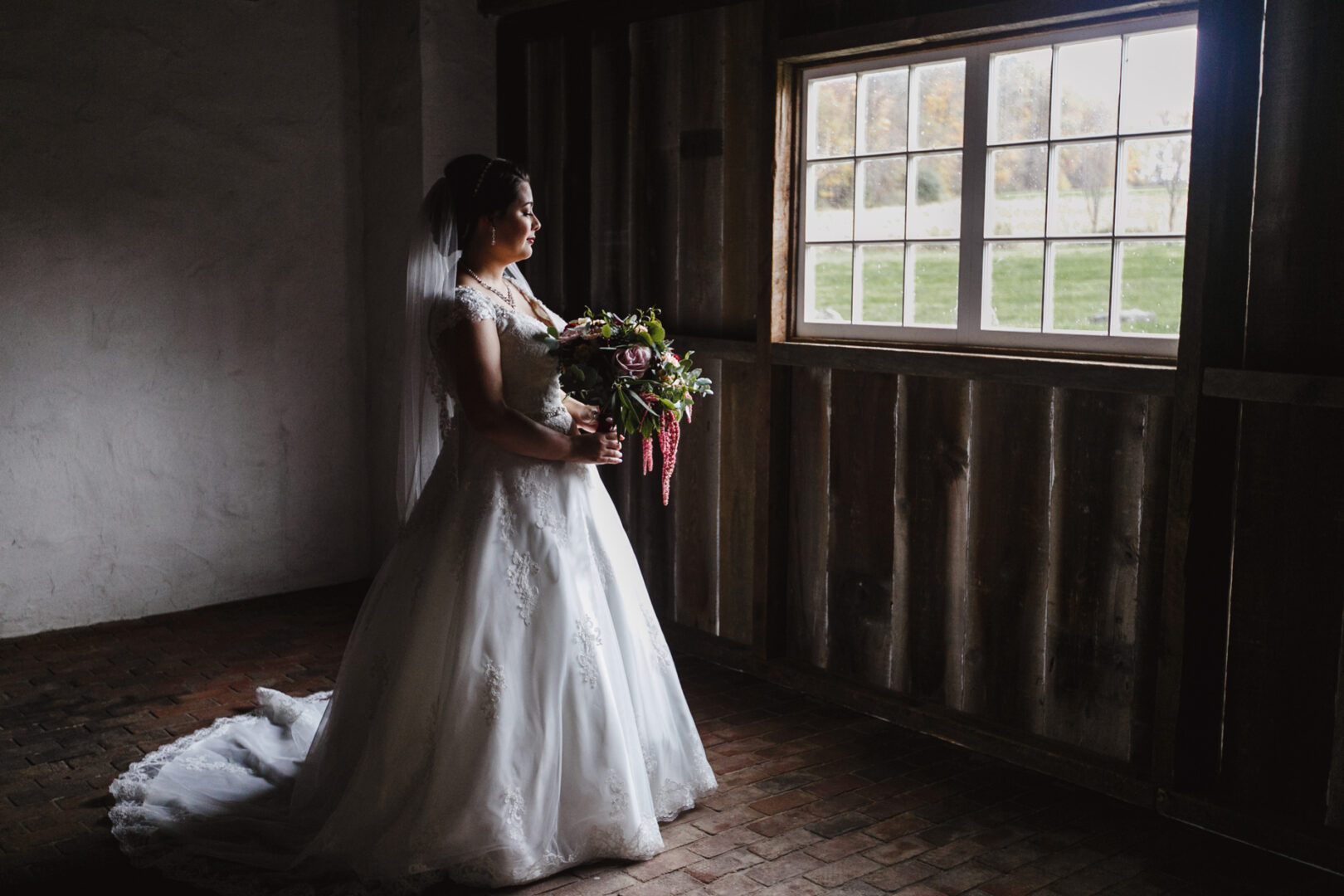 Bride in a white gown holding a bouquet stands indoors near a window with rustic wooden walls and a brick floor, gazing outside at the picturesque grounds of Brandywine Manor House.