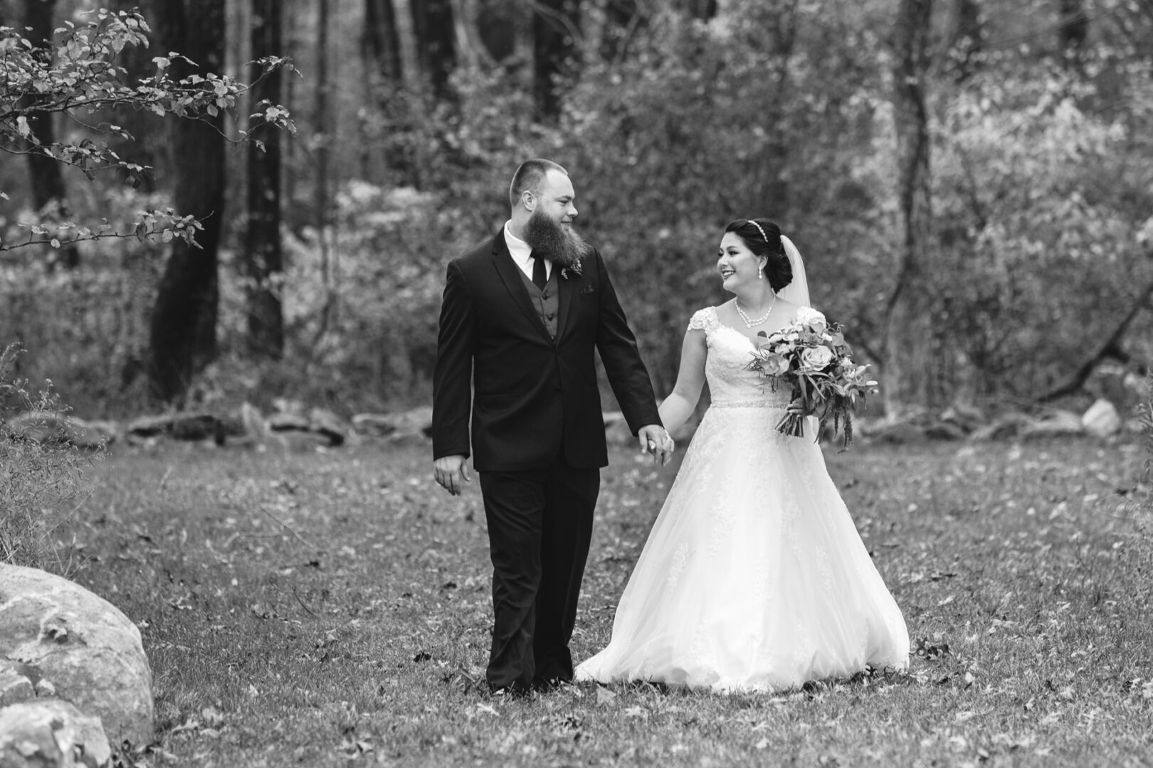 A bride and groom walk hand in hand through a wooded area at Brandywine Manor House. The bride holds a bouquet and wears a white gown, while the groom is in a dark suit, capturing the essence of an enchanting wedding day.