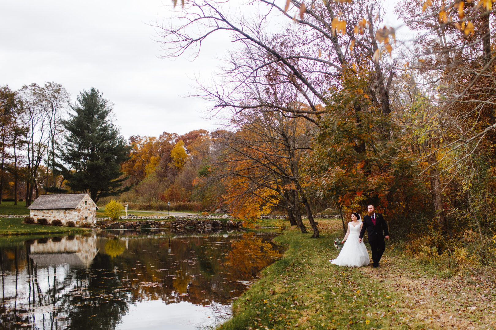 The bride and groom stroll near a serene pond surrounded by vibrant autumn foliage, with the elegant Brandywine Manor House in the background.