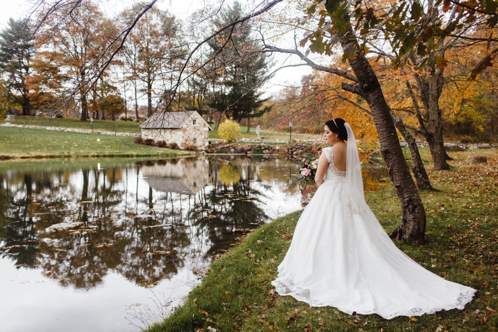 The bride, in her white wedding dress, stands gracefully by the pond at Brandywine Manor House, with autumn trees and a quaint stone building completing the picturesque backdrop.