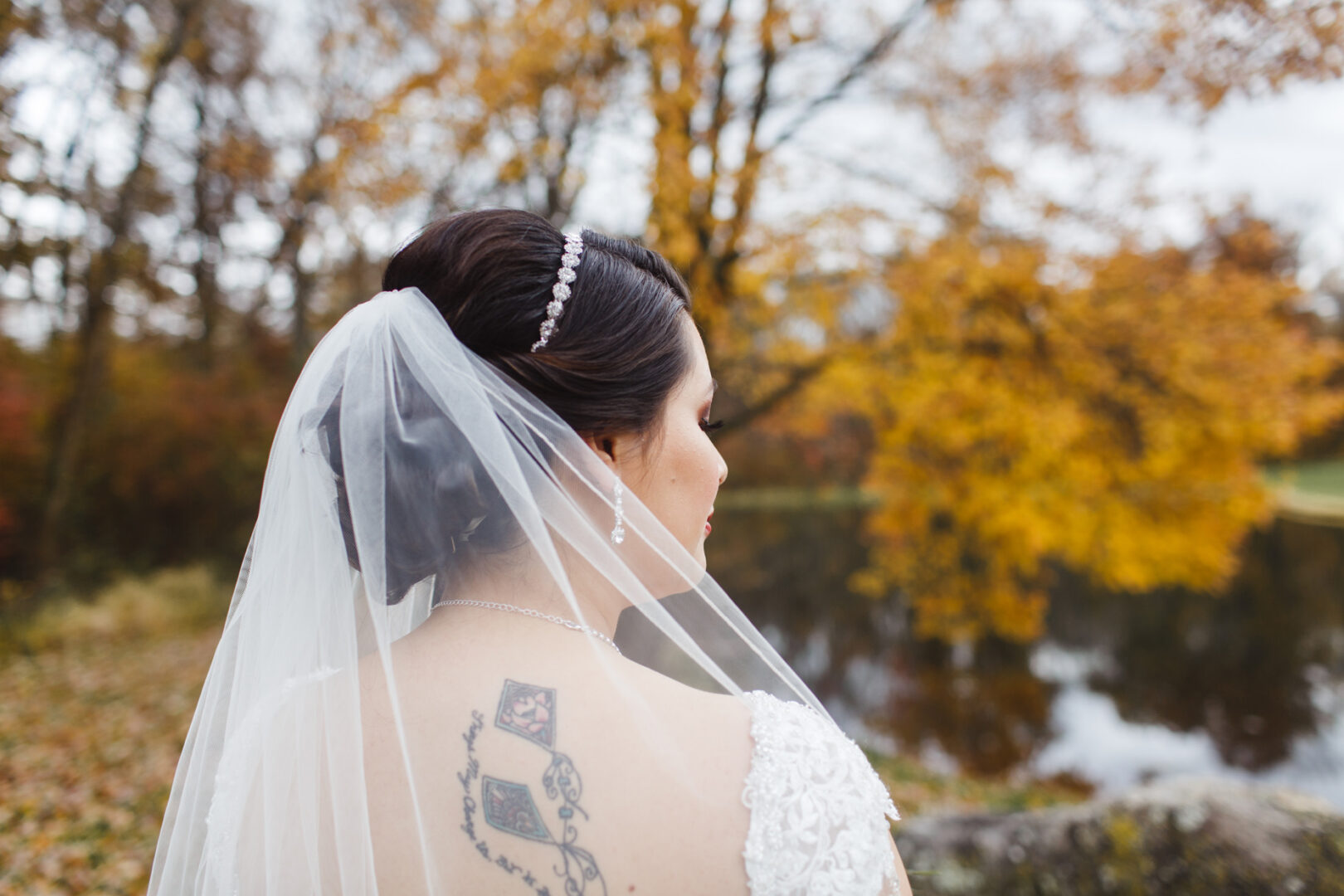 Bride standing outdoors at Brandywine Manor House, framed by autumn trees and a serene pond, wearing a veil and lace dress. Tattoo visible on shoulder.