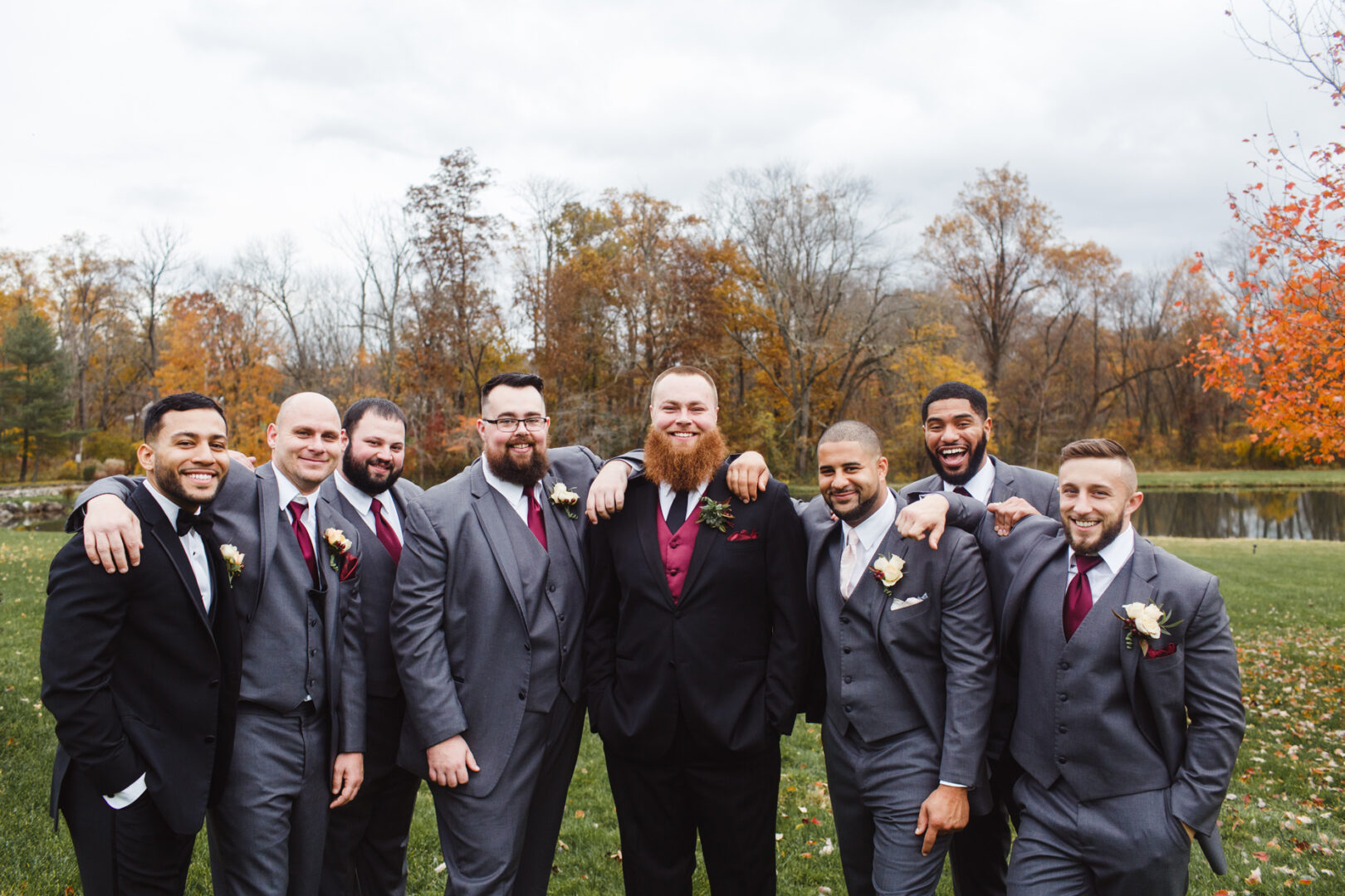 A group of nine men in suits, some wearing boutonnieres, stand together smiling in the enchanting outdoor setting of a Brandywine Manor House wedding, with lush trees providing a picturesque backdrop.