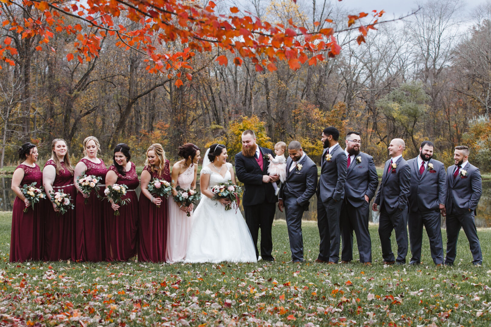 At a charming Brandywine Manor House wedding, the couple's party forms a line outdoors, bridesmaids in burgundy dresses and groomsmen in gray suits clutching bouquets. Autumn leaves gently rustle above and lay scattered across the grass, adding a rustic touch to the celebration.