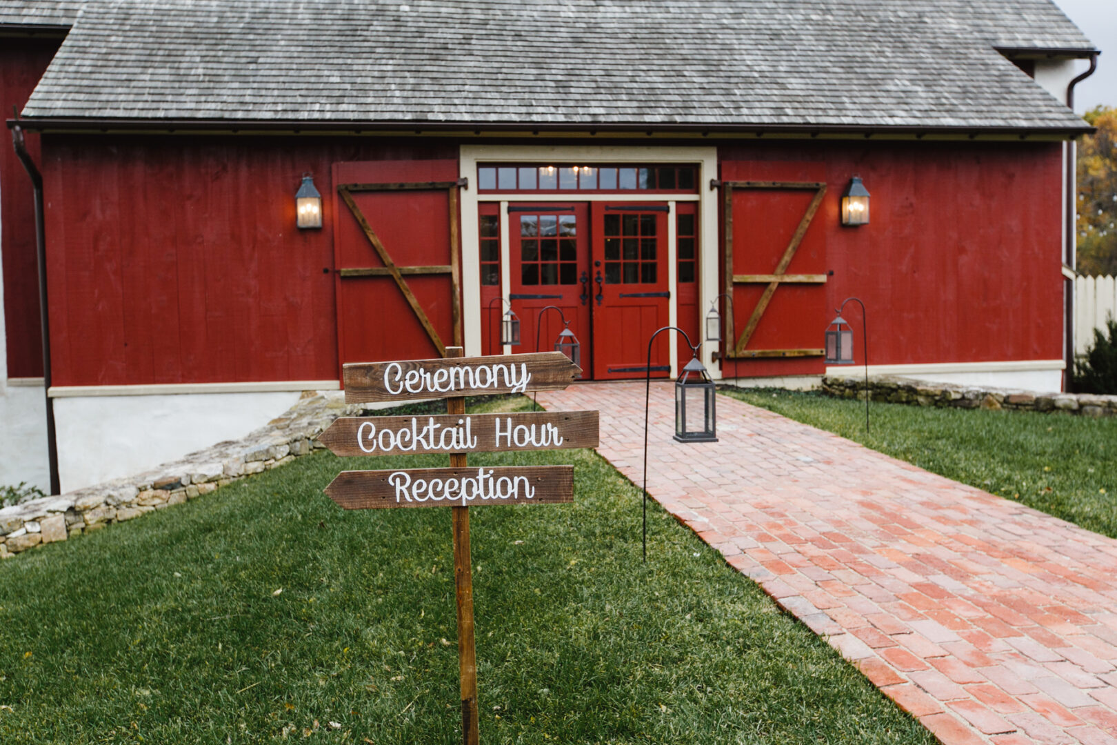 A red barn at Brandywine Manor House features a wooden signpost guiding guests to the ceremony, cocktail hour, and reception. A charming brick path leads to the entrance, flanked by lanterns creating a warm and inviting glow.