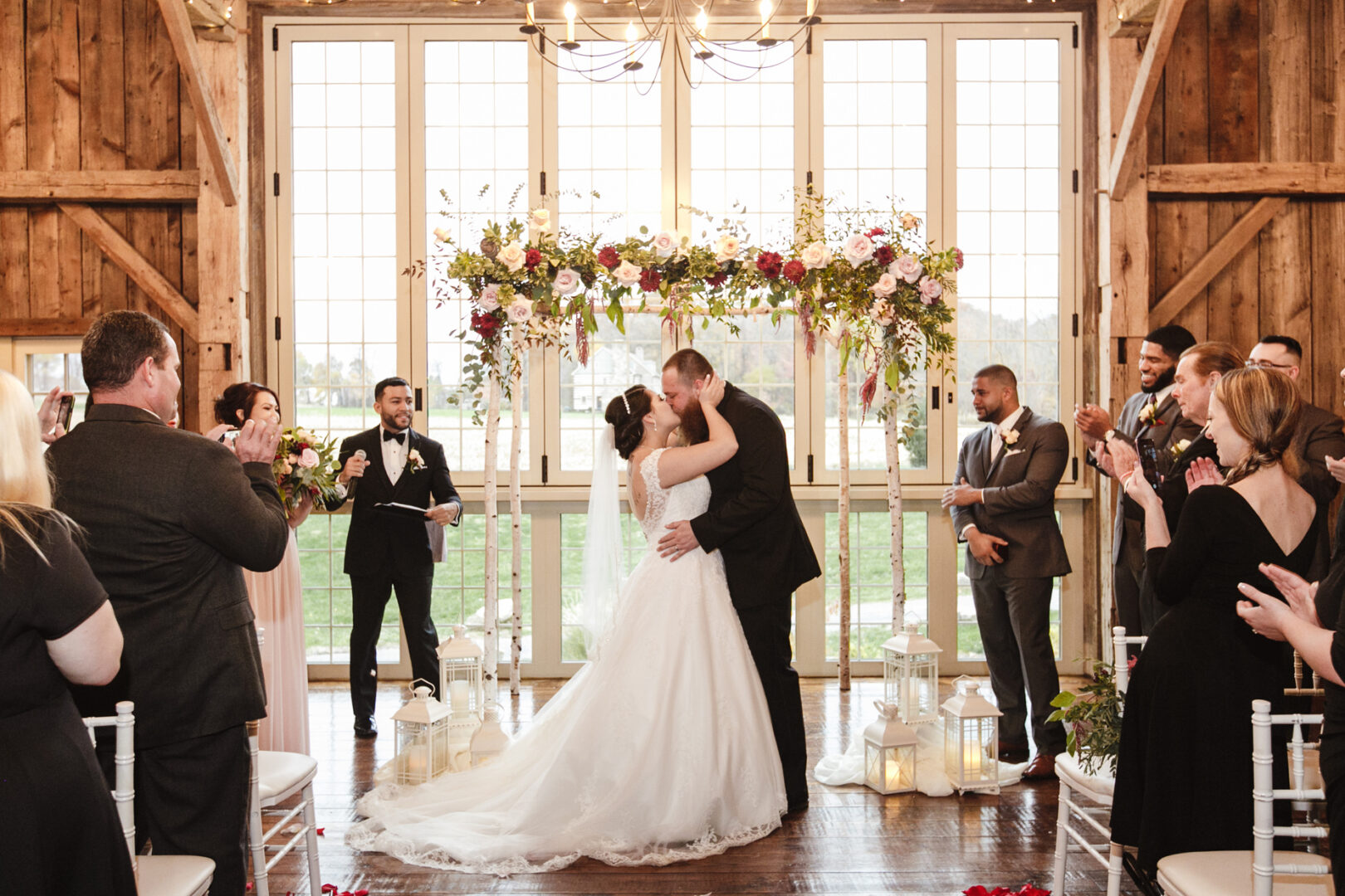 Bride and groom share a tender kiss at their Brandywine Manor House wedding, with guests clapping wildly as they stand beneath a stunning floral arch in the rustic barn setting.