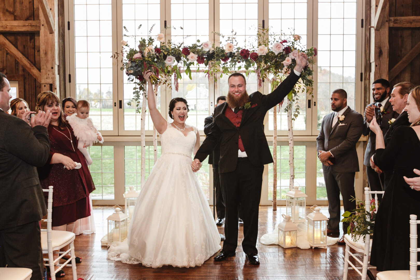 The bride and groom celebrate joyously, holding hands and raising arms, in front of a floral archway at their Brandywine Manor House wedding, with guests applauding their union.