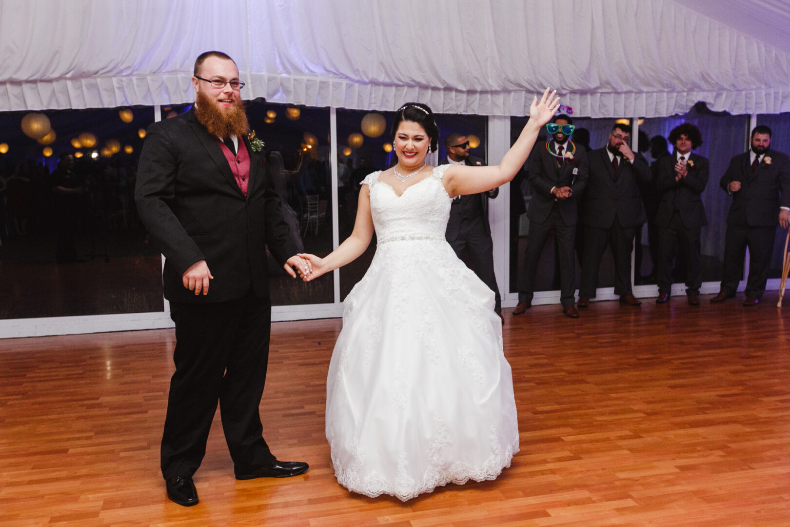 At a Brandywine Manor House wedding, the bride and groom stand on a wooden dance floor, holding hands. The bride raises one hand in celebration, while several people in formal attire stand joyfully in the background.