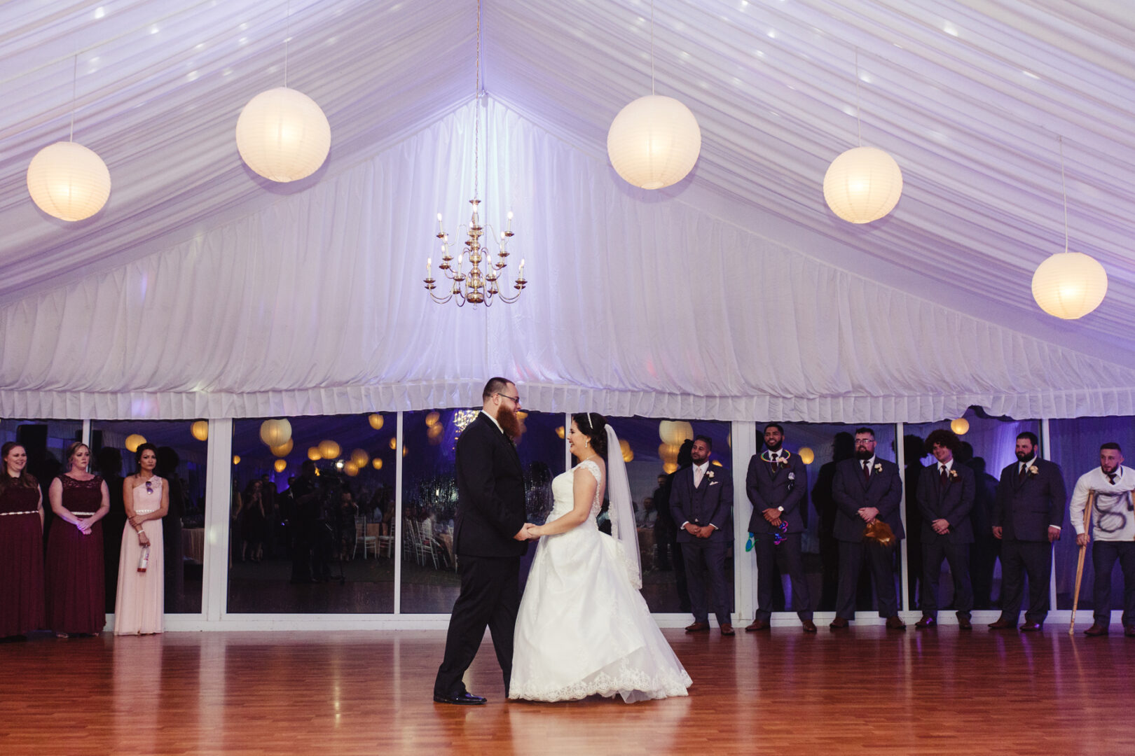 At a charming Brandywine Manor House wedding, a bride and groom gracefully dance under a tent adorned with twinkling lights, while their bridesmaids and groomsmen stand in the background, sharing in the joyous celebration.