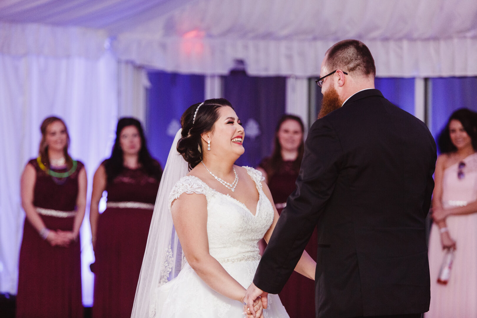 At a beautiful Brandywine Manor House wedding, the bride and groom smile warmly, holding hands as they dance. Bridesmaids look on from the background, their faces lit with joy.